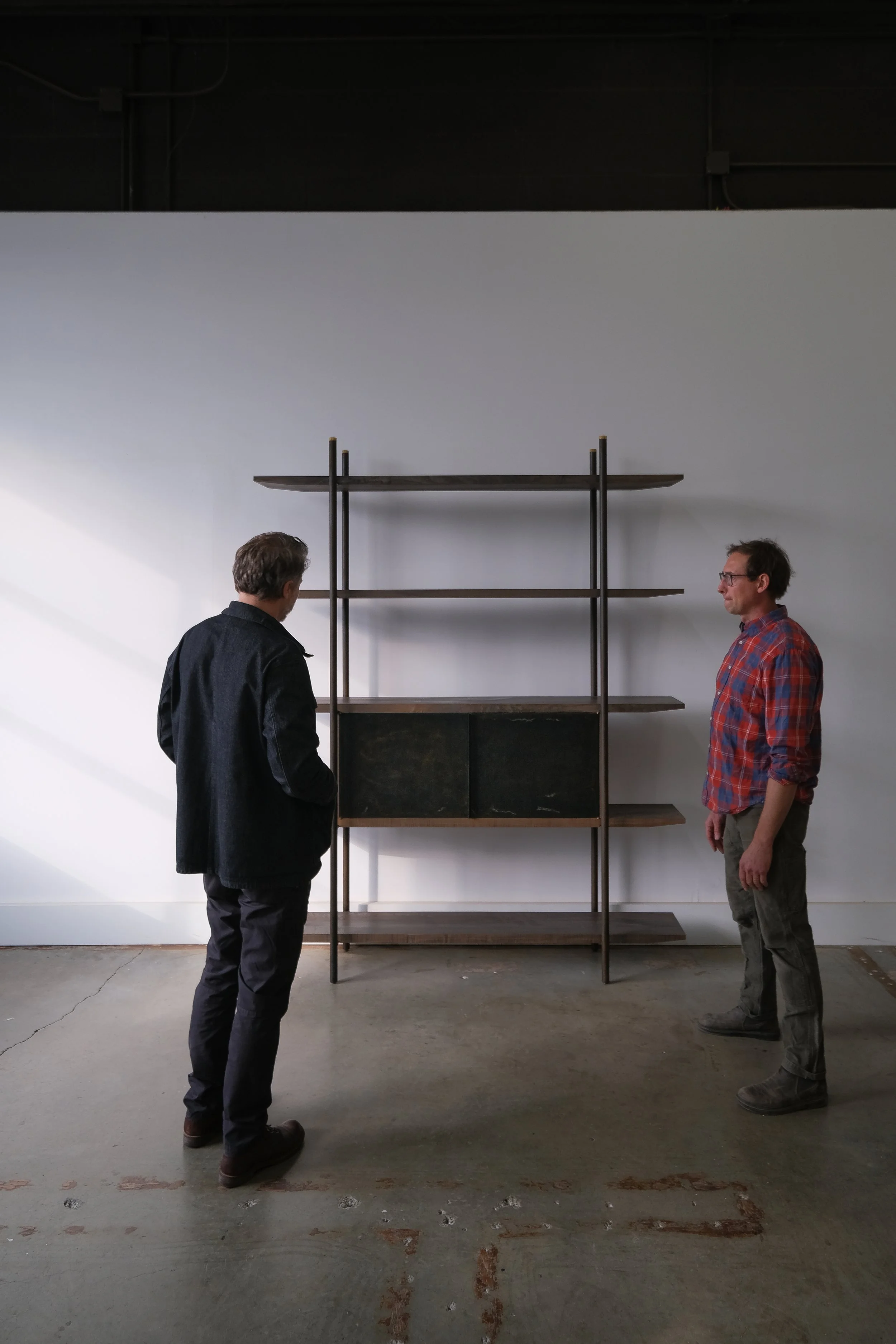 Artist Saul Becker and Chadhaus  stand facing each other in front of a minimalist wooden and metal shelving unit against a white wall.