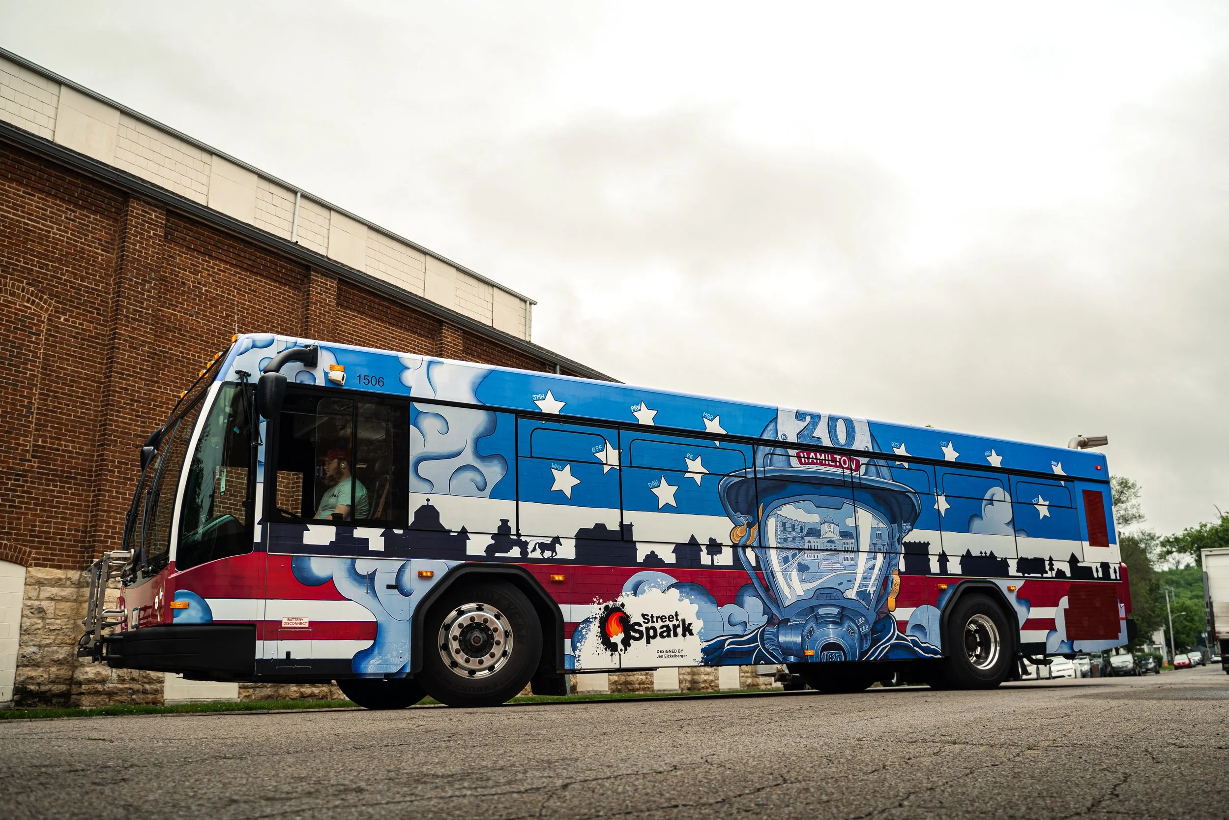 A bus painted with patriotic American flag colors and a graphic of a firefighter's helmet, with the Street Spark logo, parked on a street with a brick building and cloudy sky in the background.