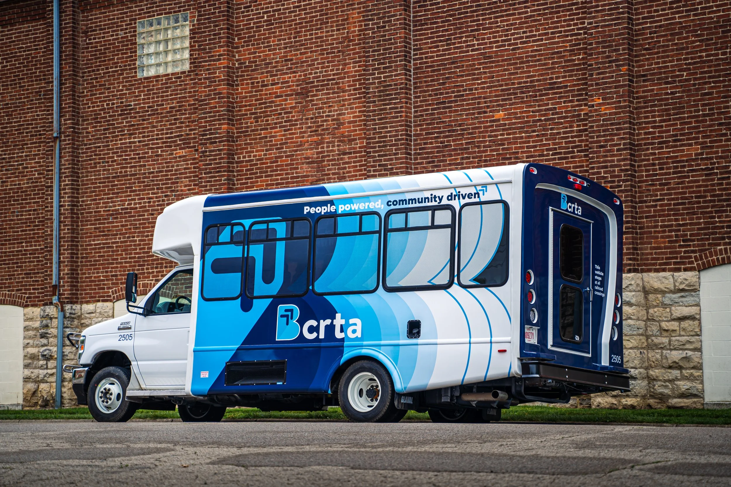 Electric bus with blue and white design, branded with BRTa and slogan 'People powered, community driven,' parked on street against brick wall.
