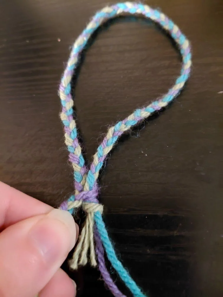 A looped braid on a dark surface, with the camera's focus on the knotted end and how the purple strand goes around it.