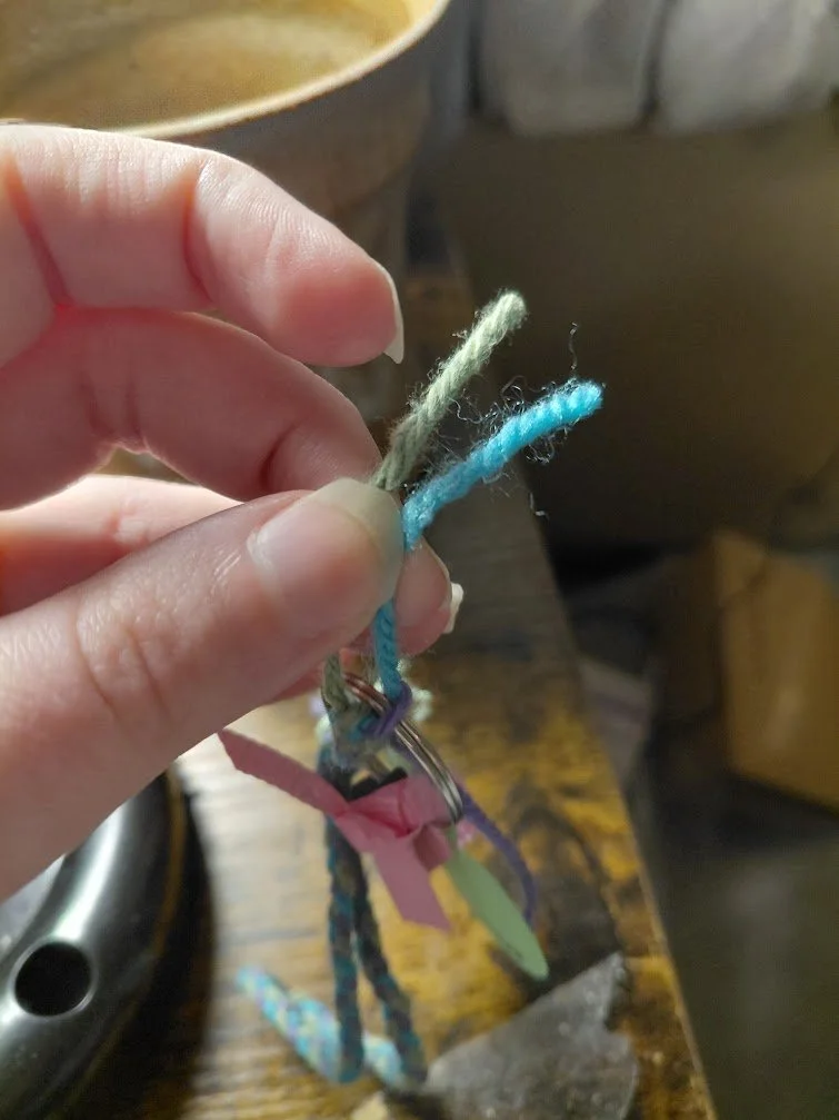 A photo of the project, being held by the green and blue strands. Below the fingers is a keyring, with the purple yarn seen looped over the keyring. the green strand is on one side of the keyring, and the blue strand on the other side.