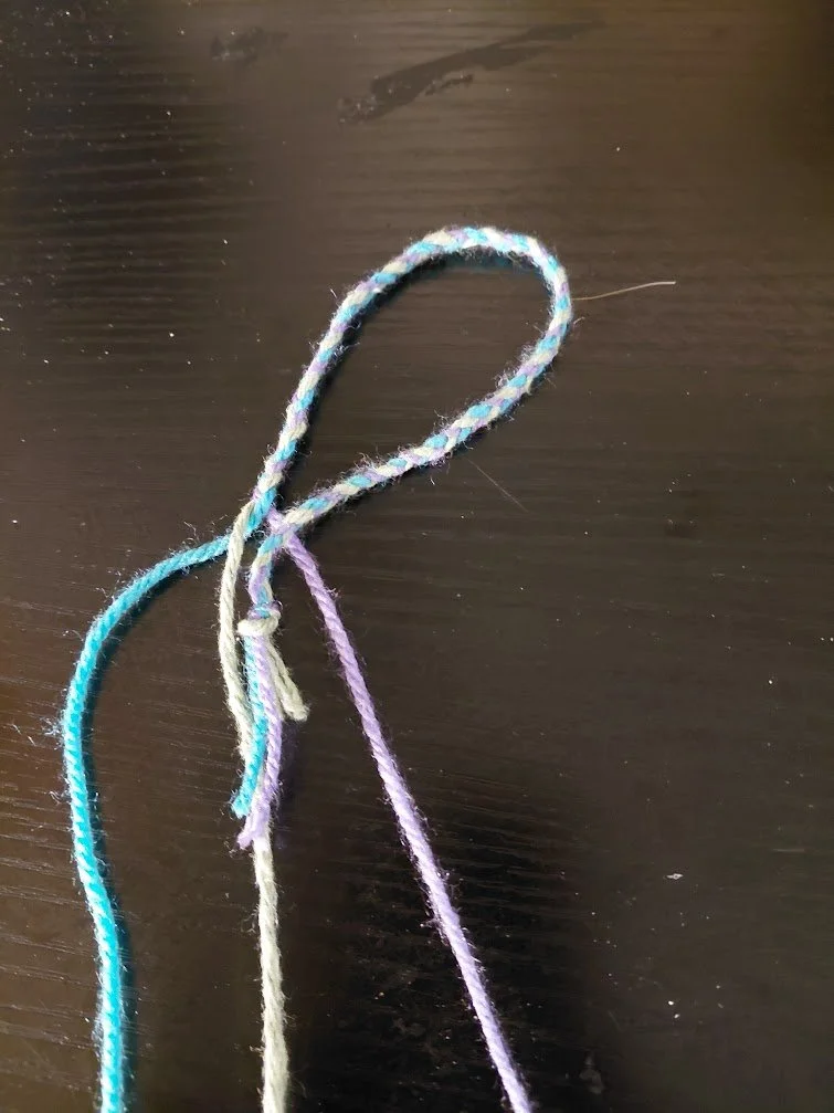 Braided yarn is now in a loop, with the knotted end in between the purple and green strands.
