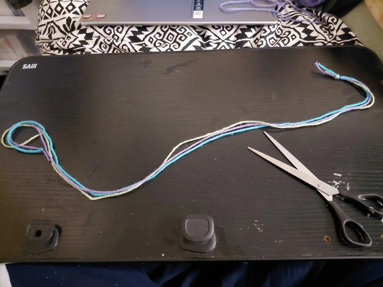 A photo of three yarn strands, knotted together on one side, laid out on a lapdesk. A pair of scissors is artfully to the side.