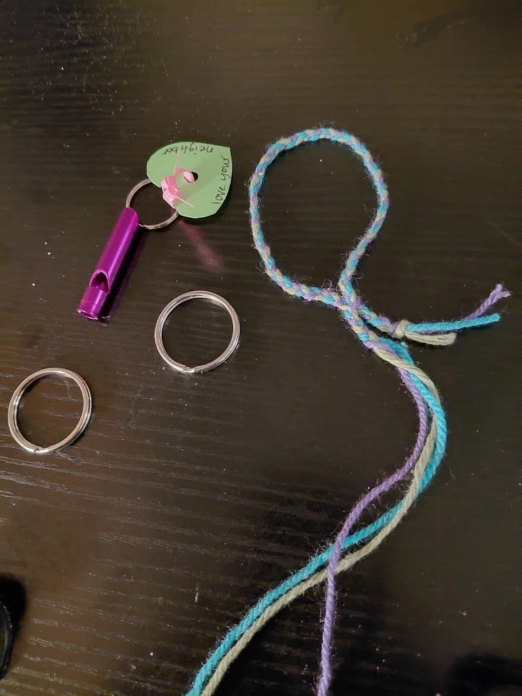 A braided loop, with an unfinished end, and the finished end looped in the braid.  Also on the dark surface are a couple of keey wrings, and a purple whistle already with key ring though has a tag that says, "Love your neighbor"