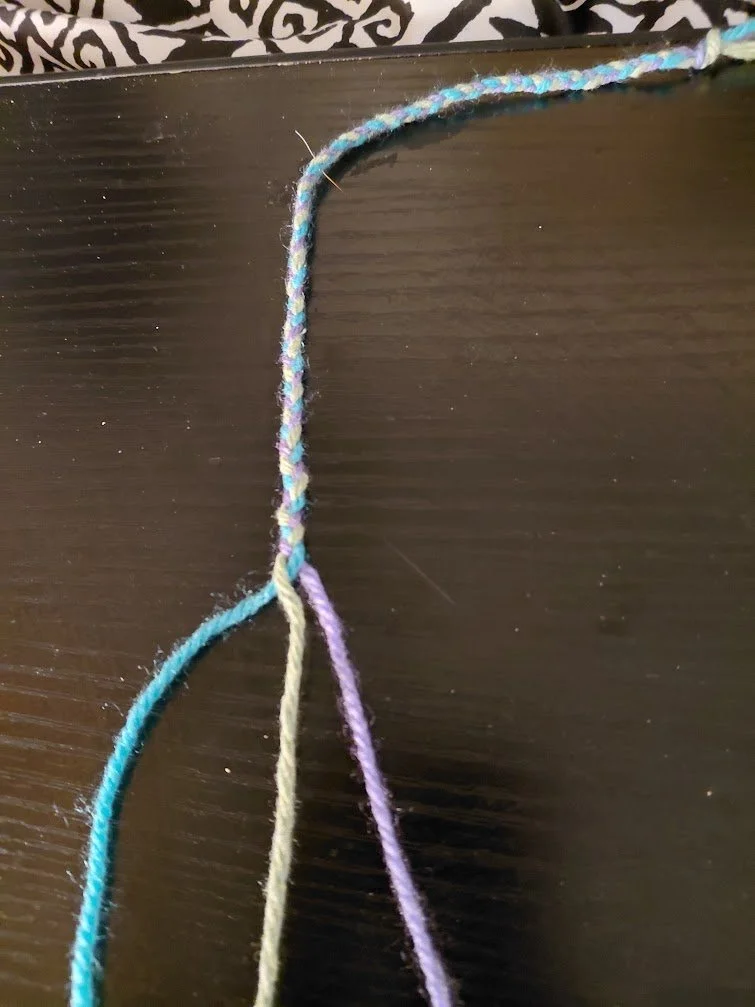 Braided yarn, vertically laid out on a dark surface. The strands are separated out at the bottom with blue on the left, green in the center, purple on the right.