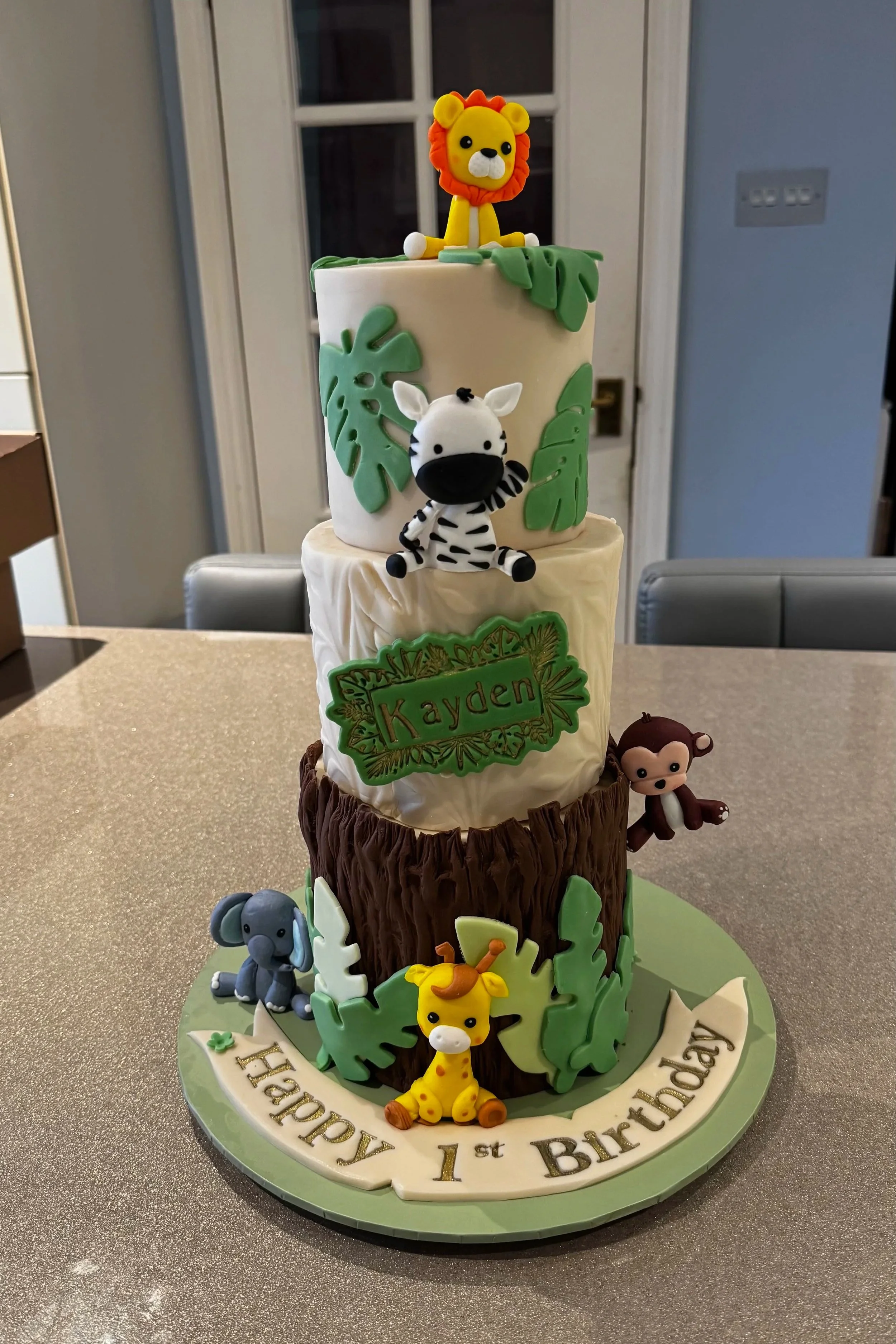 Three tier, jungle themed celebration cake, with shades of green foliage, resin animal characters, wording on plaque and cake board.