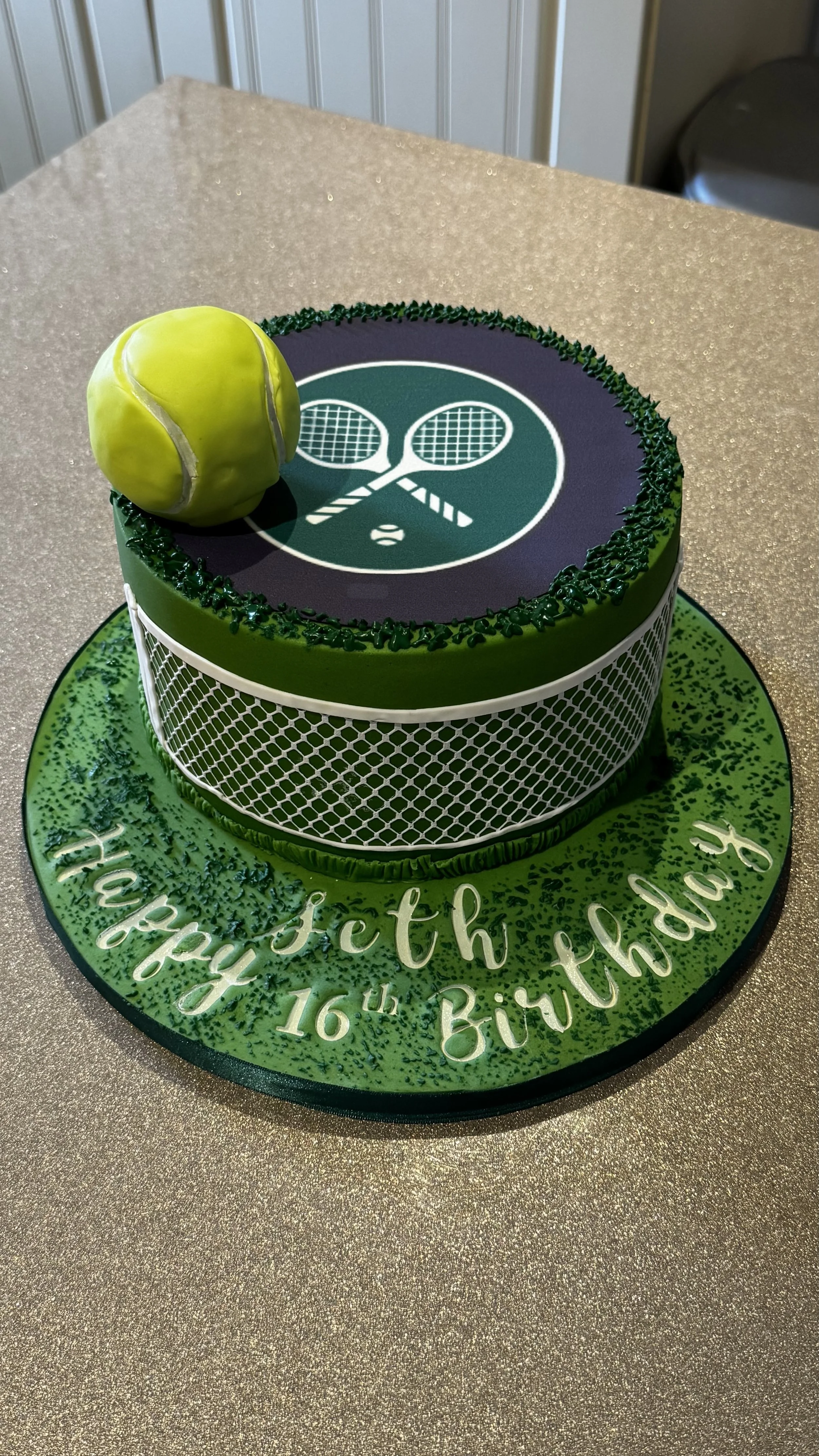 A birthday cake decorated with a tennis theme, including a tennis ball and tennis racket images on the top and net design on the sides, with a green and purple  colour scheme, and the message on the base.