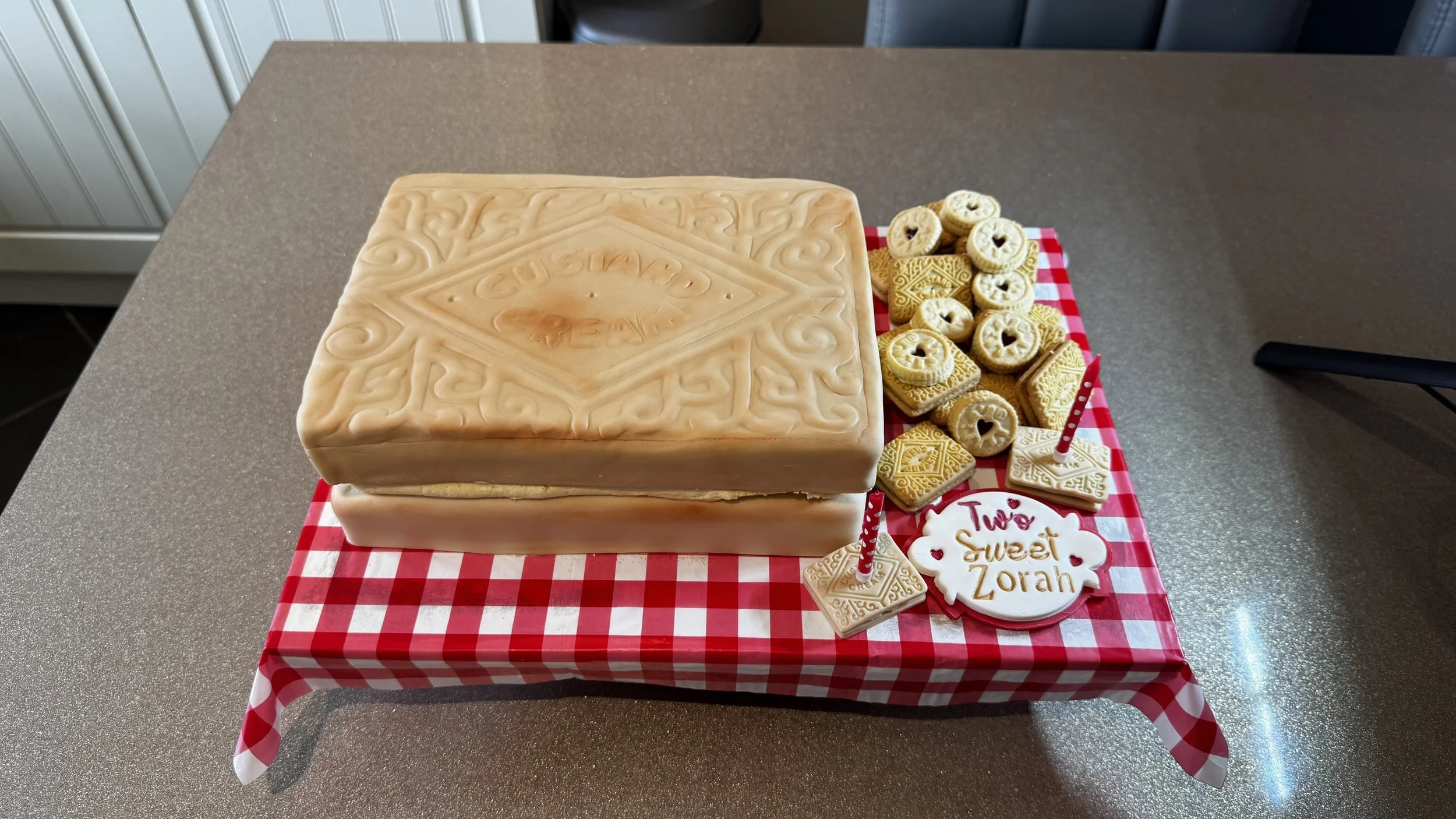A large custard cream biscuit cake shaped surrounded by assorted cookies with heart-shaped cutouts and decorative patterns, placed on a red and white checkered tablecloth.