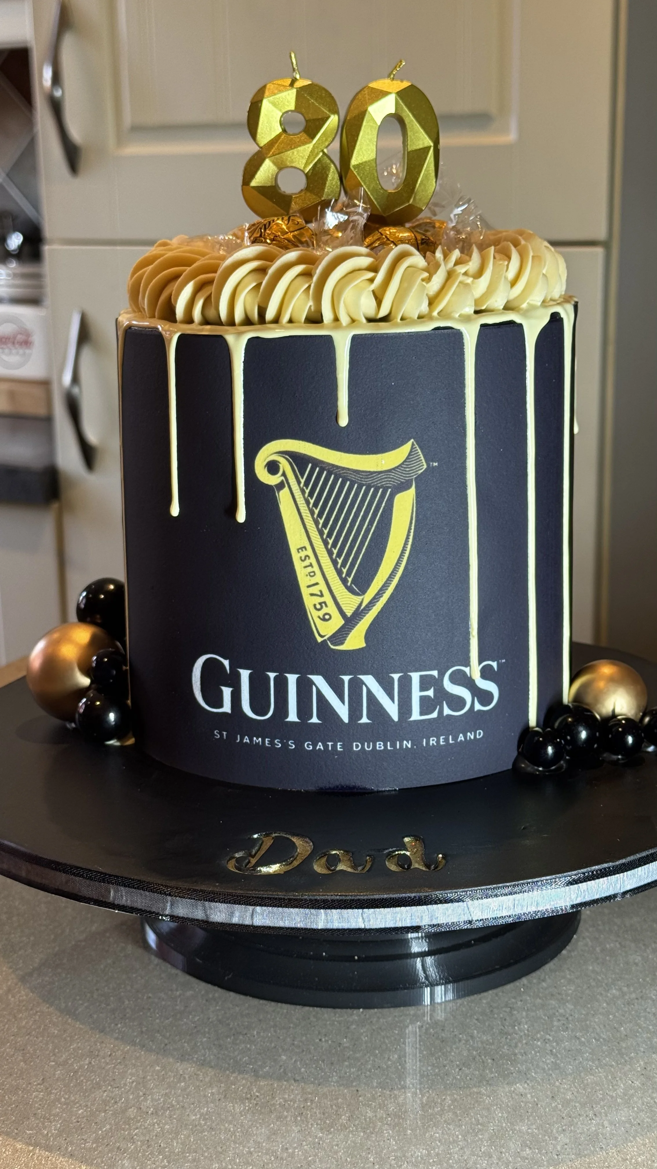 Black and gold  cake with a harp logo, gold and black decorative balls, and gold '80' birthday candles on top, placed on a black cake board with a gold 'Dad' inscription.