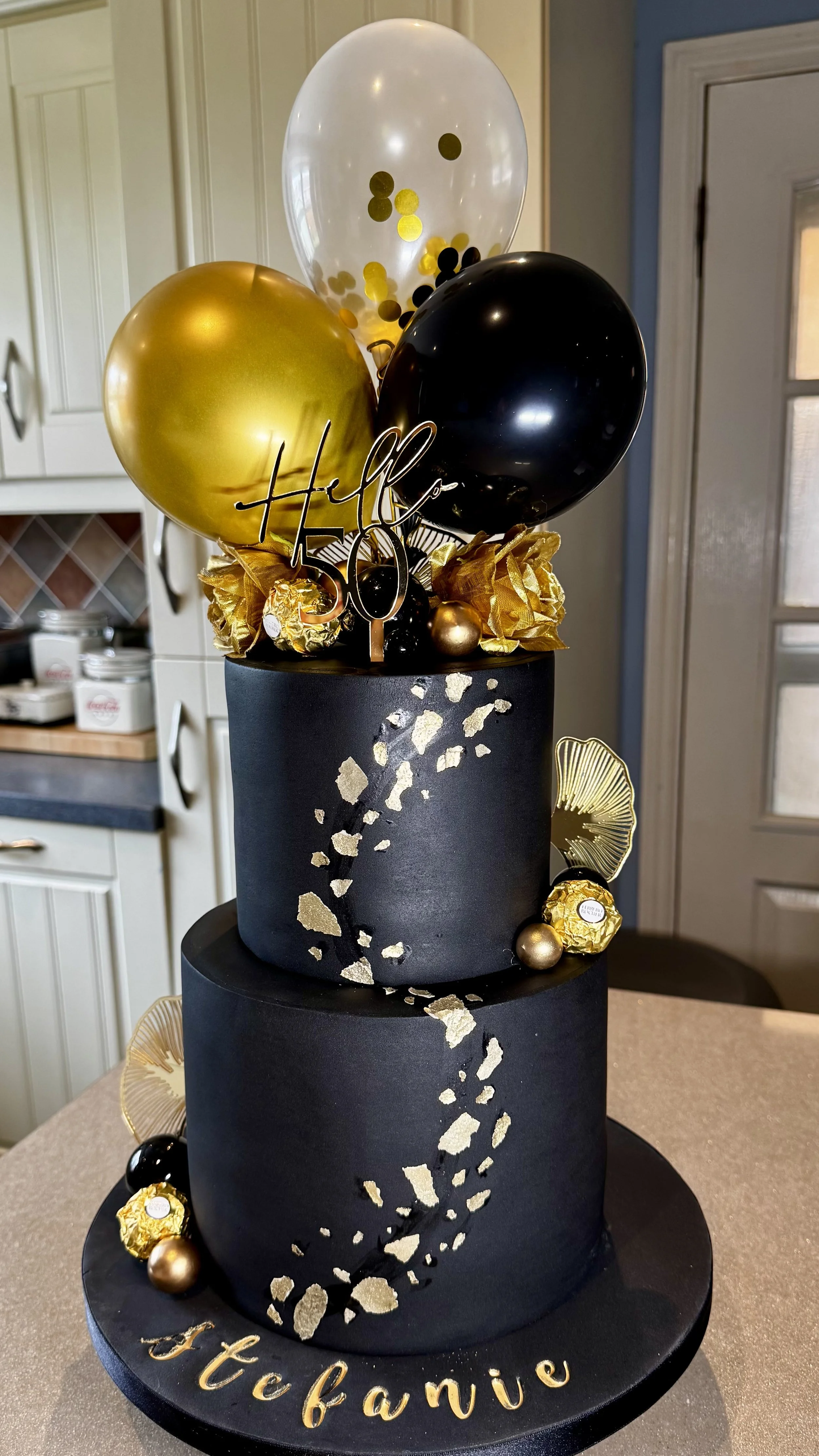 Black and Gold two tier celebration cake with gold acrylic topper and 5 inch balloons.
