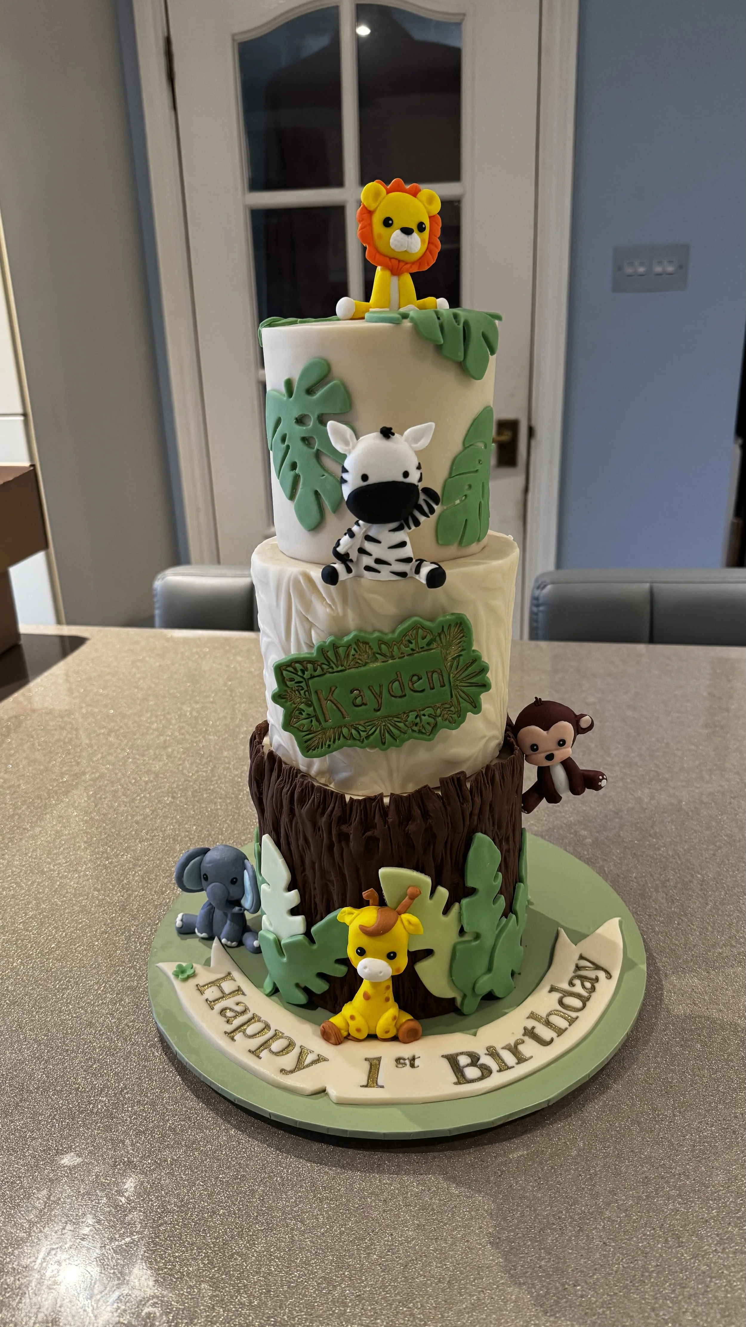 Three tier, jungle themed celebration cake, with shades of green foliage, resin animal characters, wording on plaque and cake board.
