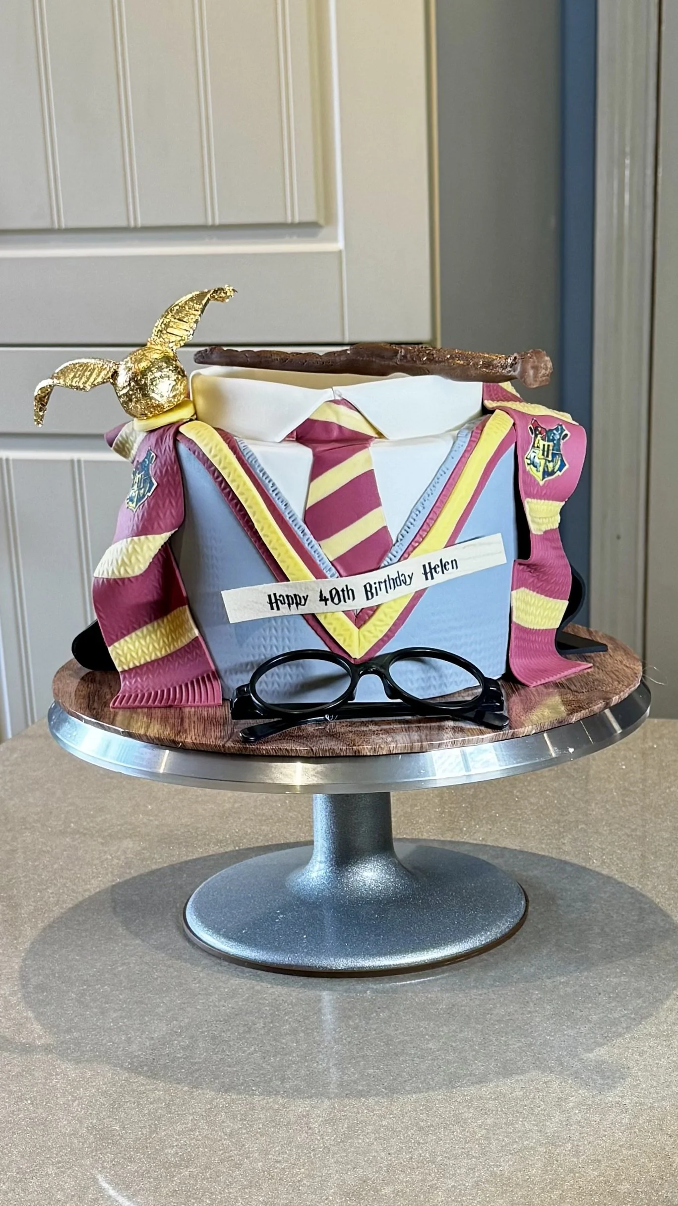 A birthday cake decorated with a Harry Potter theme, including a Gryffindor scarf, glasses, and a broomstick, with a gold-hatched chocolate frog on top, and a message reading 'Happy 40th Birthday Helen'.