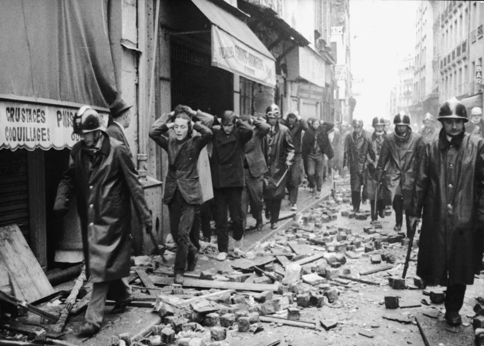 Students, flanked by police in helmets and long coats, walk through a rubble-strewn street with their hands on the backs of their heads, black and white image