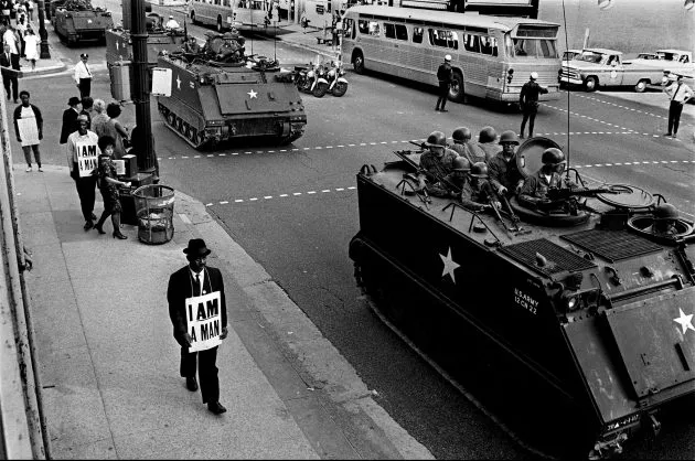 A black and white image shows a man in an hat and suit marching next to a tank, wearing a sign that reads I AM A MAN