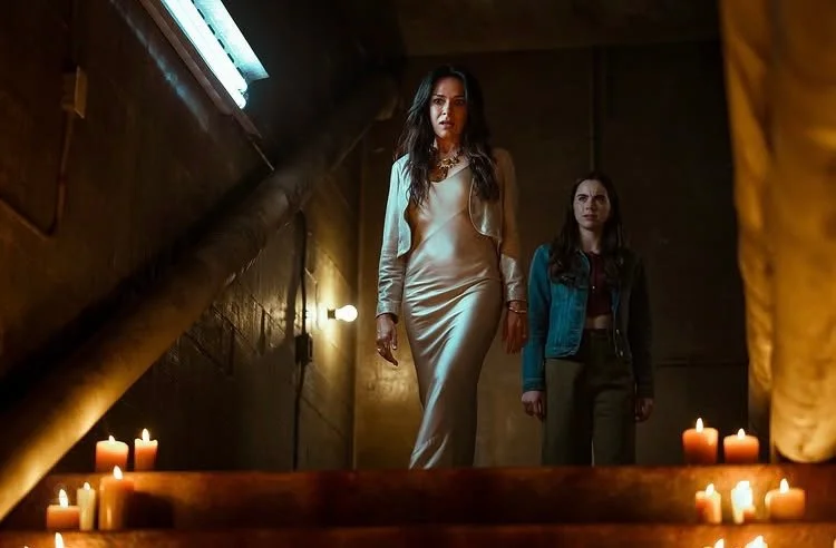 A slender woman with long dark hair wearing a beige bias-cut silk gown and matching jacket approaches a staircase lined with candles. A young girl with long dark hair stands behind her wearing a blue jean jacket and dark green trousers.