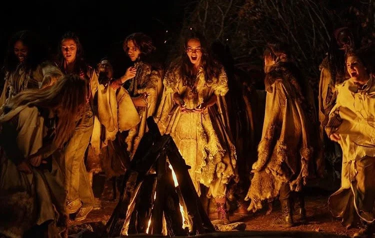 A group of young people, most apparently women and primarily light-skinned with the exception of one Black person in the upper left, wear roughly sewn leather and fur garments while dancing around a bonfire