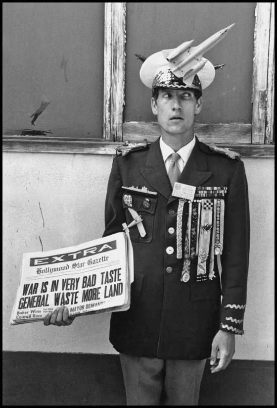 Black and white: a man in an army uniform faces the camera, his eyes glancing to the right, a model rocket attached to his hat, holding a fake newspaper with a headline reading WAR IS IN VERY BAD TASTE GENERAL WASTE MORE LAND