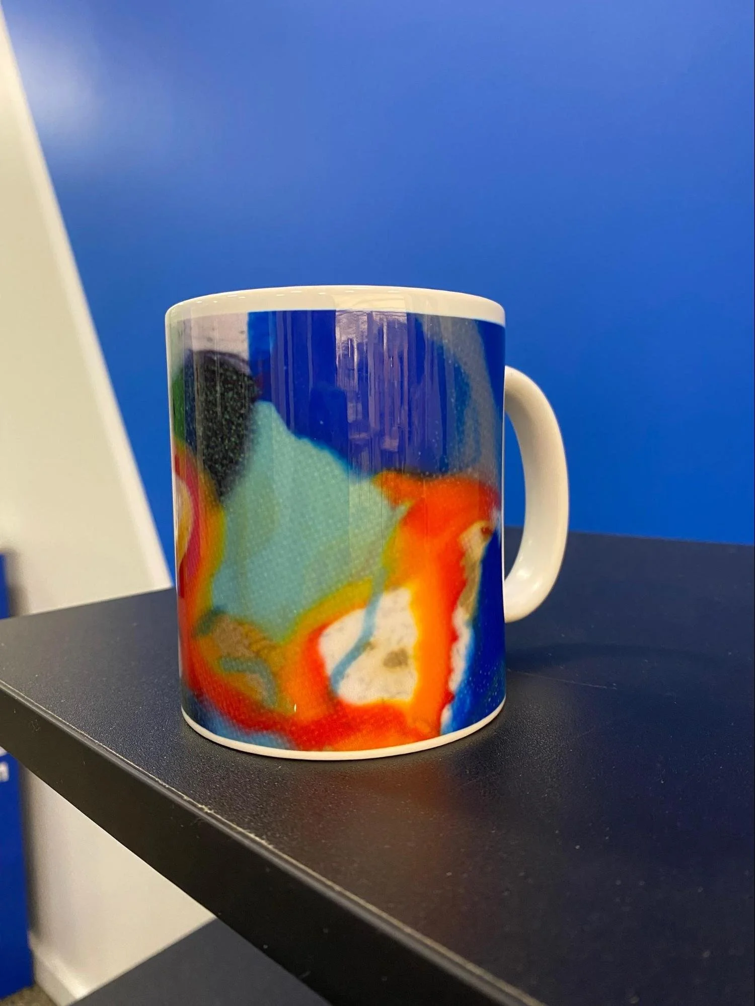 a ceramic mug printed with an abstract pattern in cobalt and turquoise, yellow, red, orange, black and white sits on the corner of a black table. The wall behind is a lighter blue, with a stripe of white emerging diagonally from the upper left corner