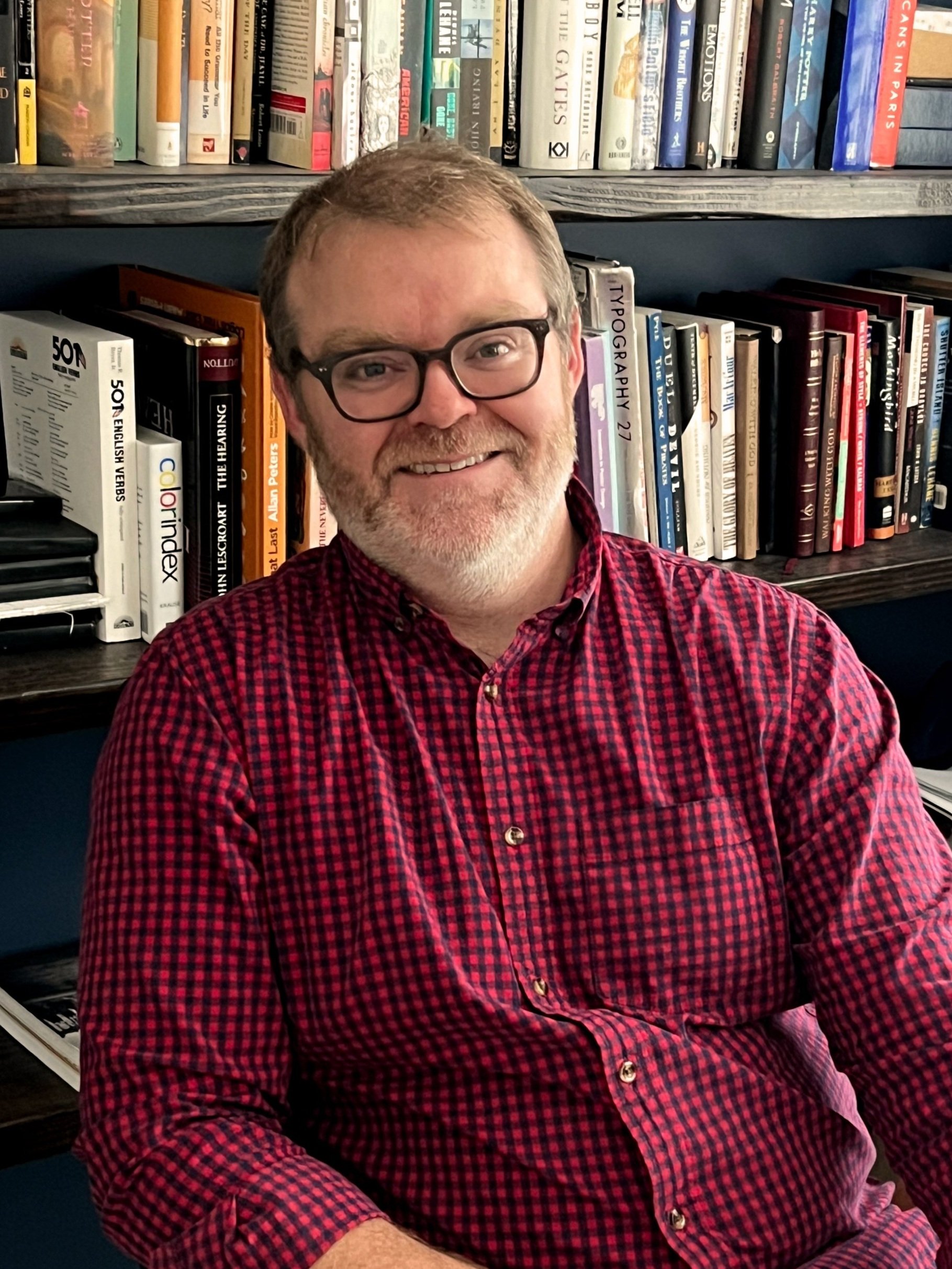 Person in a checkered shirt sitting in front of bookshelves filled with various books.