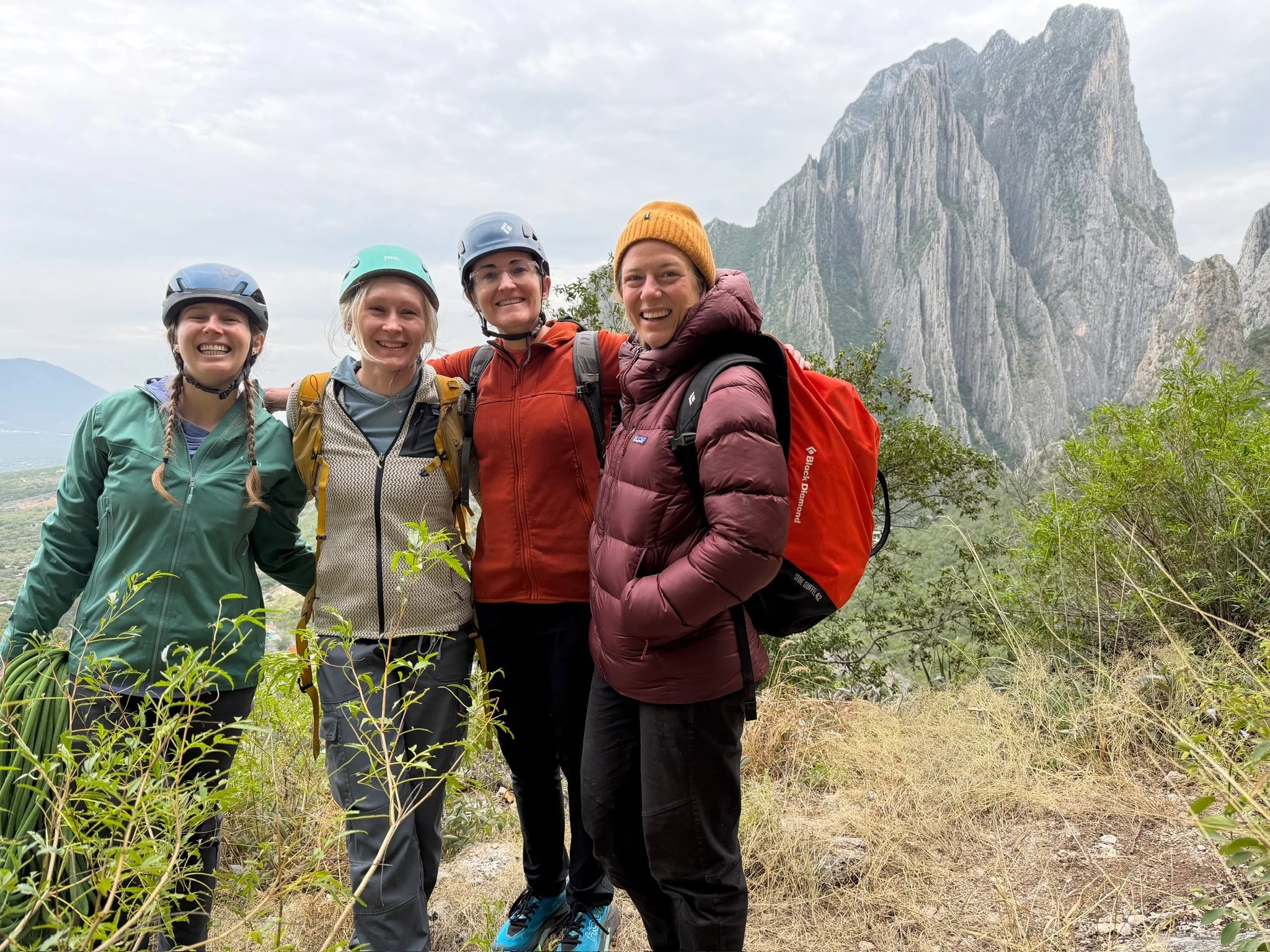 Four women in outdoor hiking gear smiling with arms around each other, standing on a trail with mountains and cloudy sky in the background.