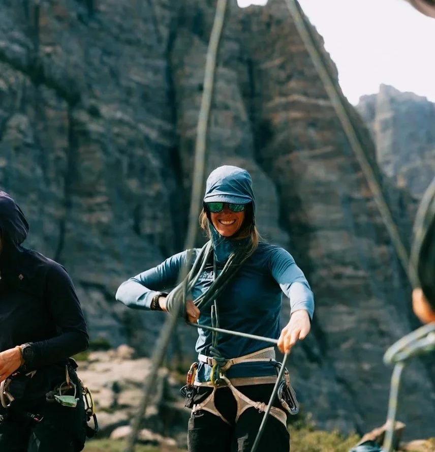 A woman in outdoor climbing gear smiling outdoors with a rocky cliff in the background.