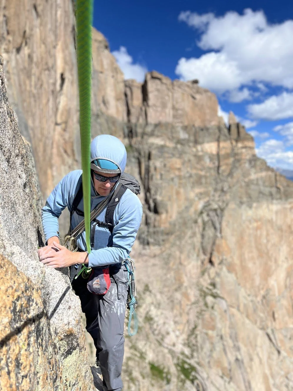 A male rock climber wearing a blue hoodie, gray pants, sunglasses, and a harness is ascending a steep rock face with a green climbing rope. The background features a rocky mountain landscape under a blue sky with scattered clouds.