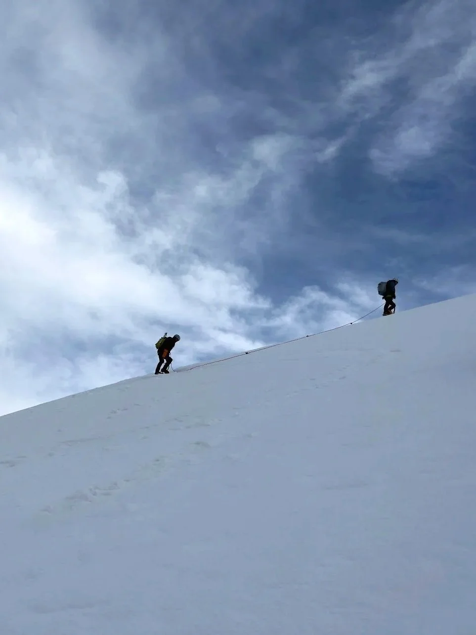 Two hikers climbing a snowy mountain under a cloudy sky.
