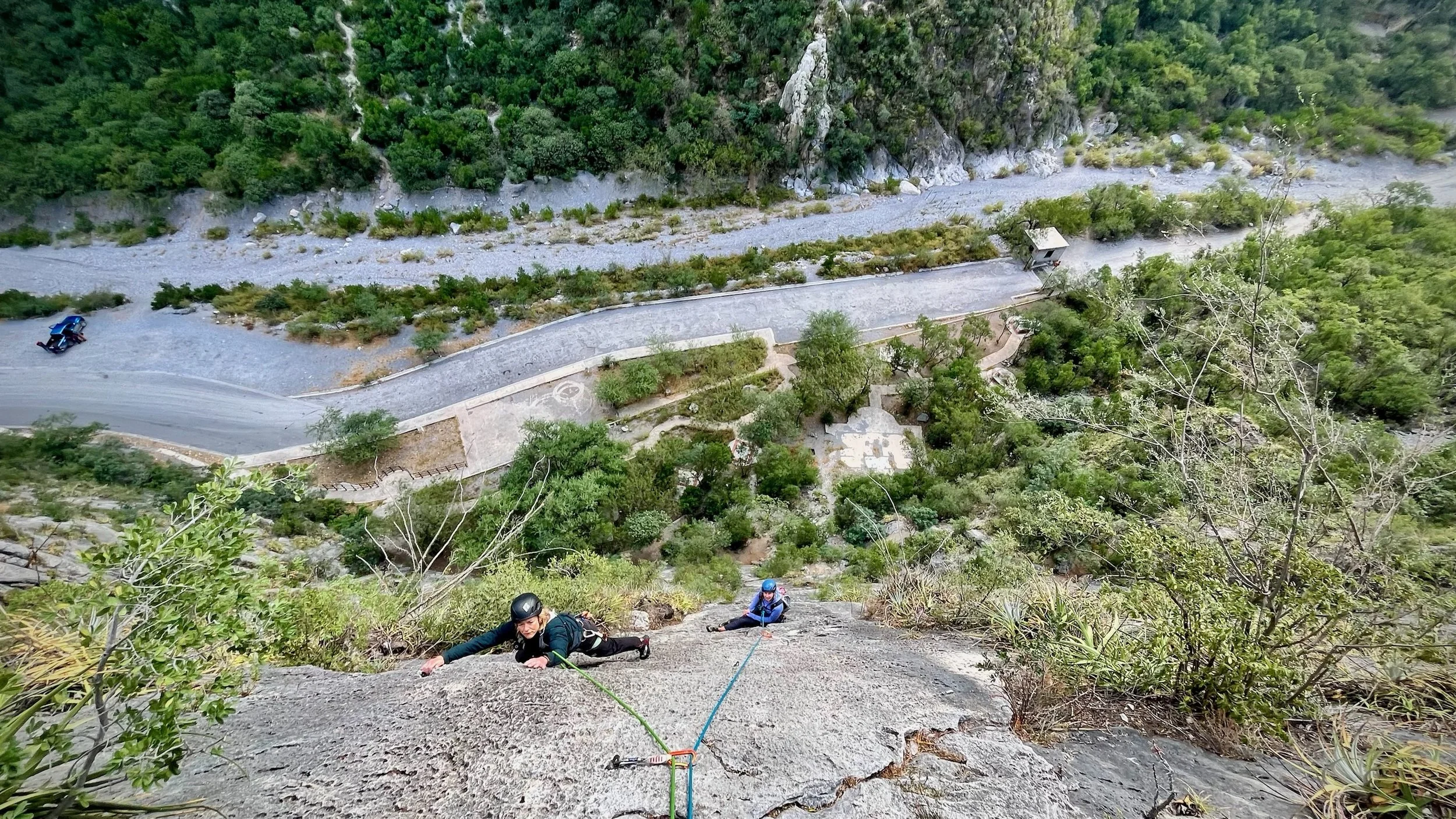 Two rock climbers ascend a steep cliff face with climbing gear, overlooking a winding road, trees, rocks, and parked vehicles in a mountainous area.