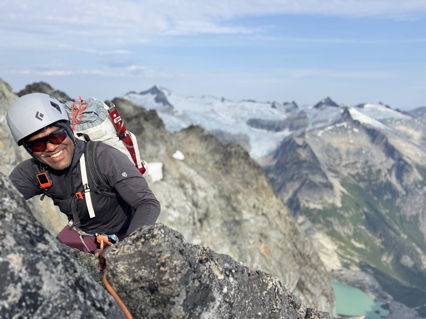 Man rock climbing on a mountain with snowy peaks in the background, wearing a helmet, sunglasses, and outdoor gear.