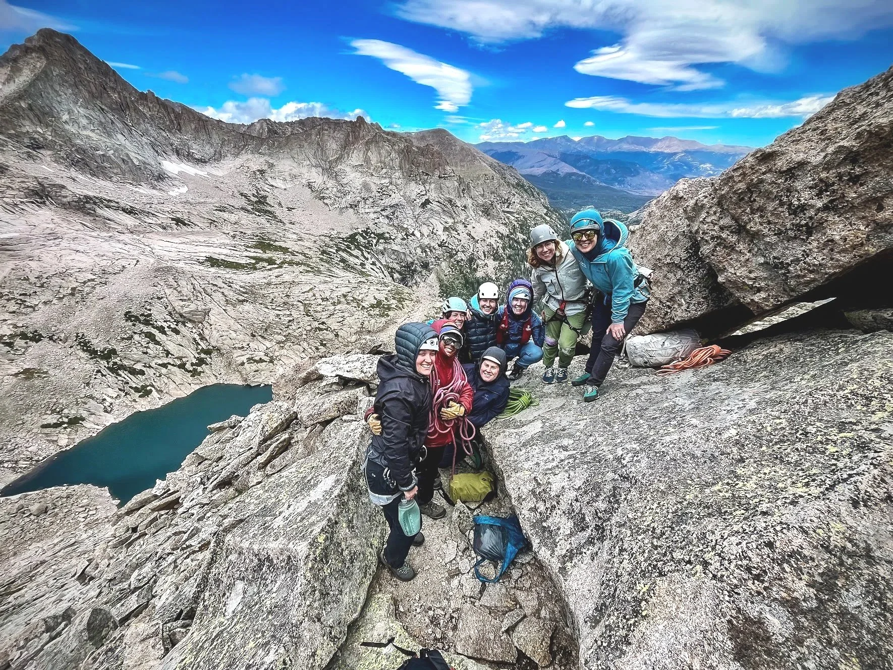 Group of climbers or hikers on a rocky mountain ledge with a scenic mountain landscape and blue sky in the background.