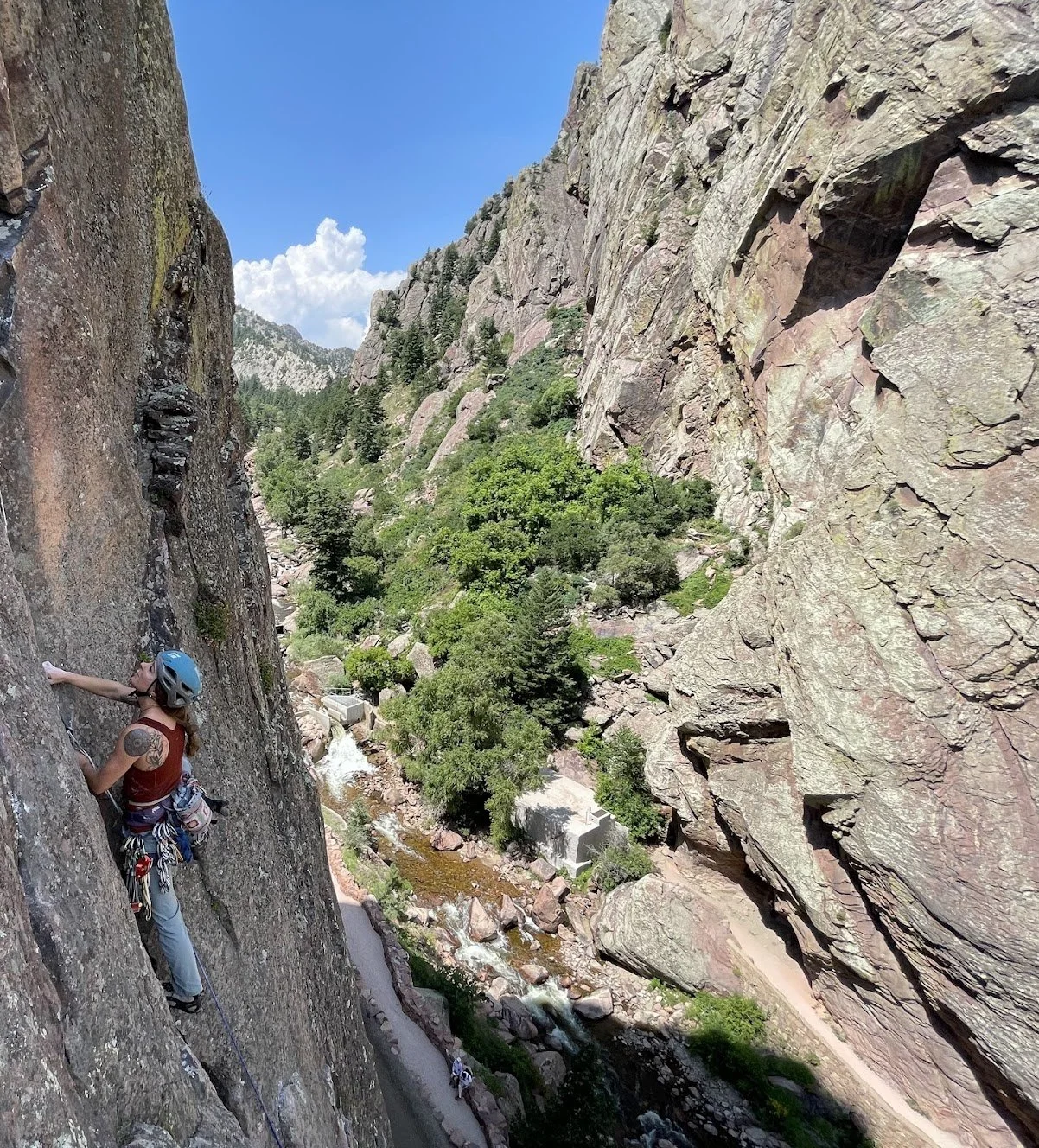 A woman rock climbing on a steep cliffside with a blue helmet, situated in a mountainous area with green trees and a river below under a blue sky.