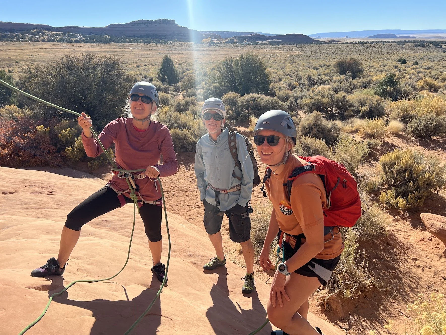 Three people wearing helmets and sunglasses, with one holding a climbing rope, standing on a rocky desert landscape with shrubs and distant mesas under a clear blue sky.