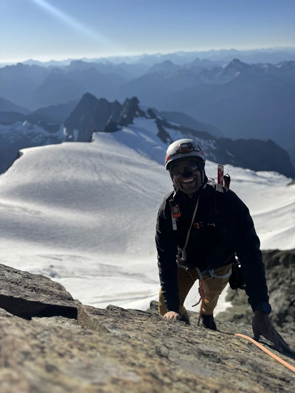A male climber wearing a helmet, sunglasses, and climbing gear scales a rocky surface on a snowy mountain, with snow-capped peaks and a distant mountain range in the background.