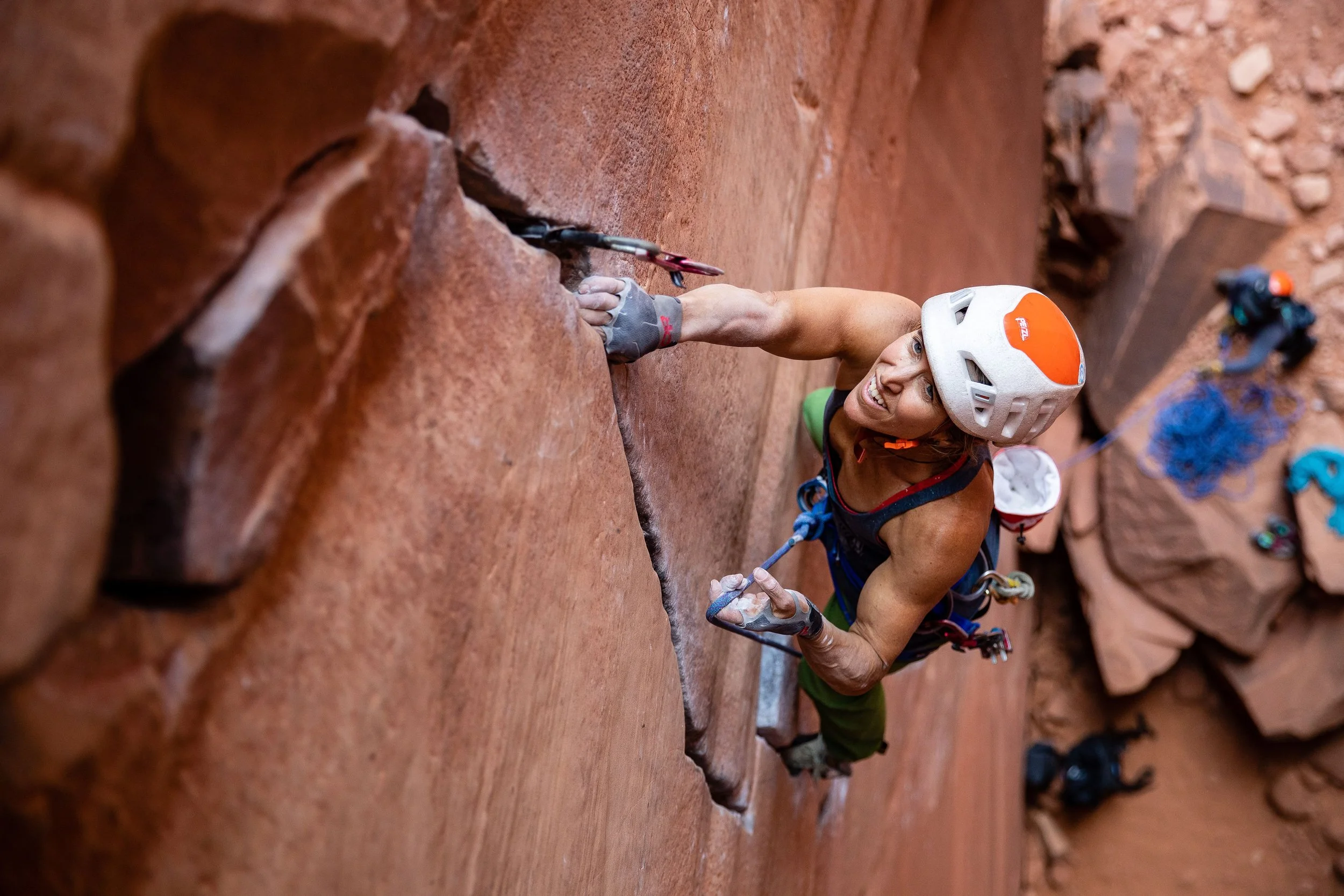A woman rock climbing on a red sandstone wall, wearing a white helmet, gloves, and athletic clothing, smiling while holding onto a climbing hold.
