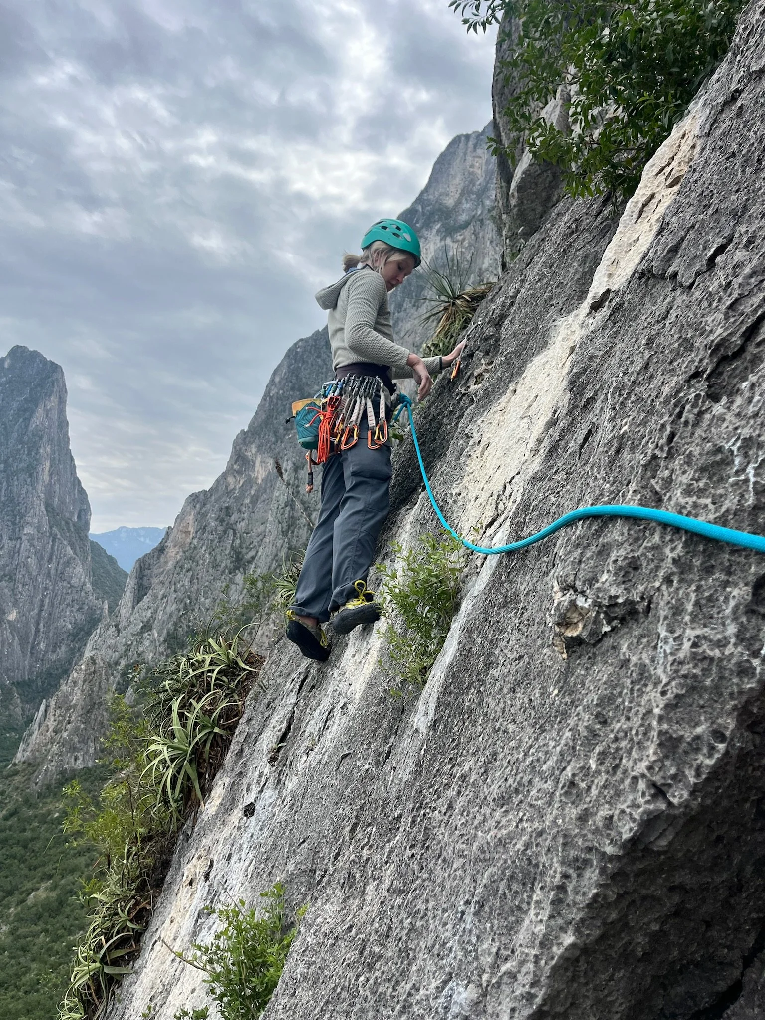 A woman climbing a rock face outdoors with climbing gear and a helmet, mountain landscape in the background.