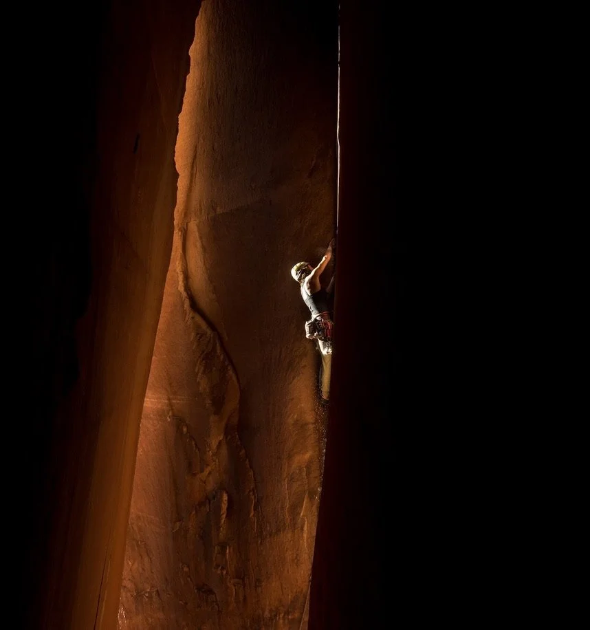 A person rock climbing in a narrow canyon with towering, reddish-brown walls.