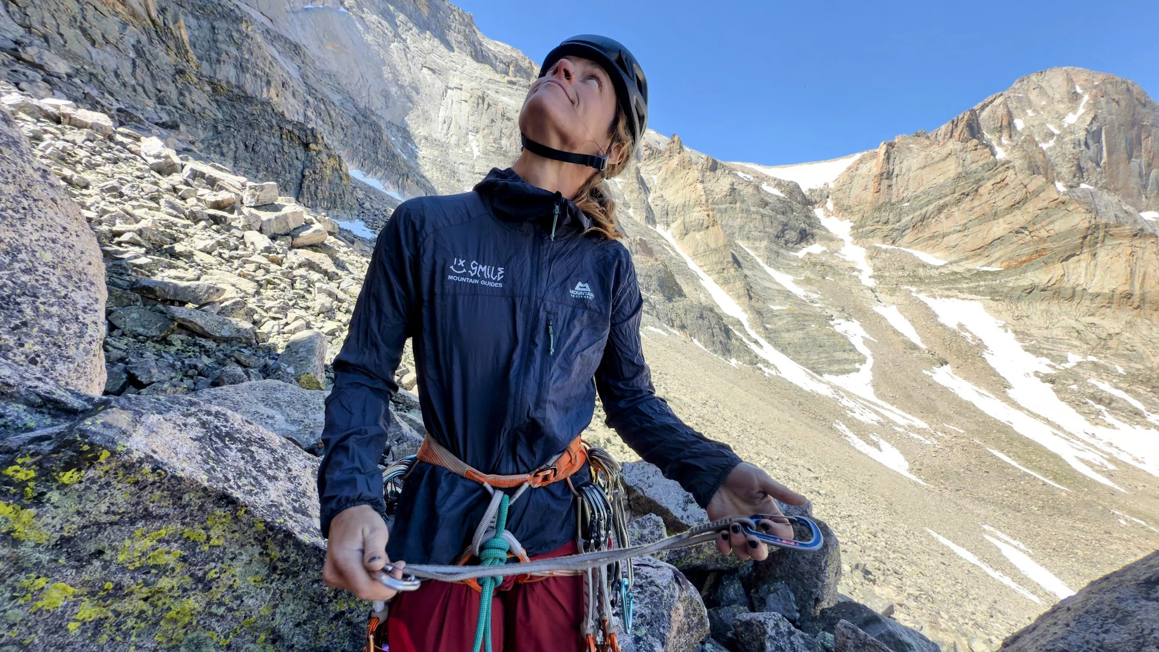 A smiling woman wearing an orange outdoor jacket and black backpack standing in a mountainous landscape with a cloudy sky in the background.