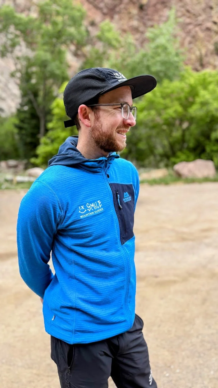 A smiling man wearing a light blue Arcteryx jacket with logos, a white and blue helmet, glasses, and an orange helmet strap, standing outdoors in a snowy, mountainous landscape.