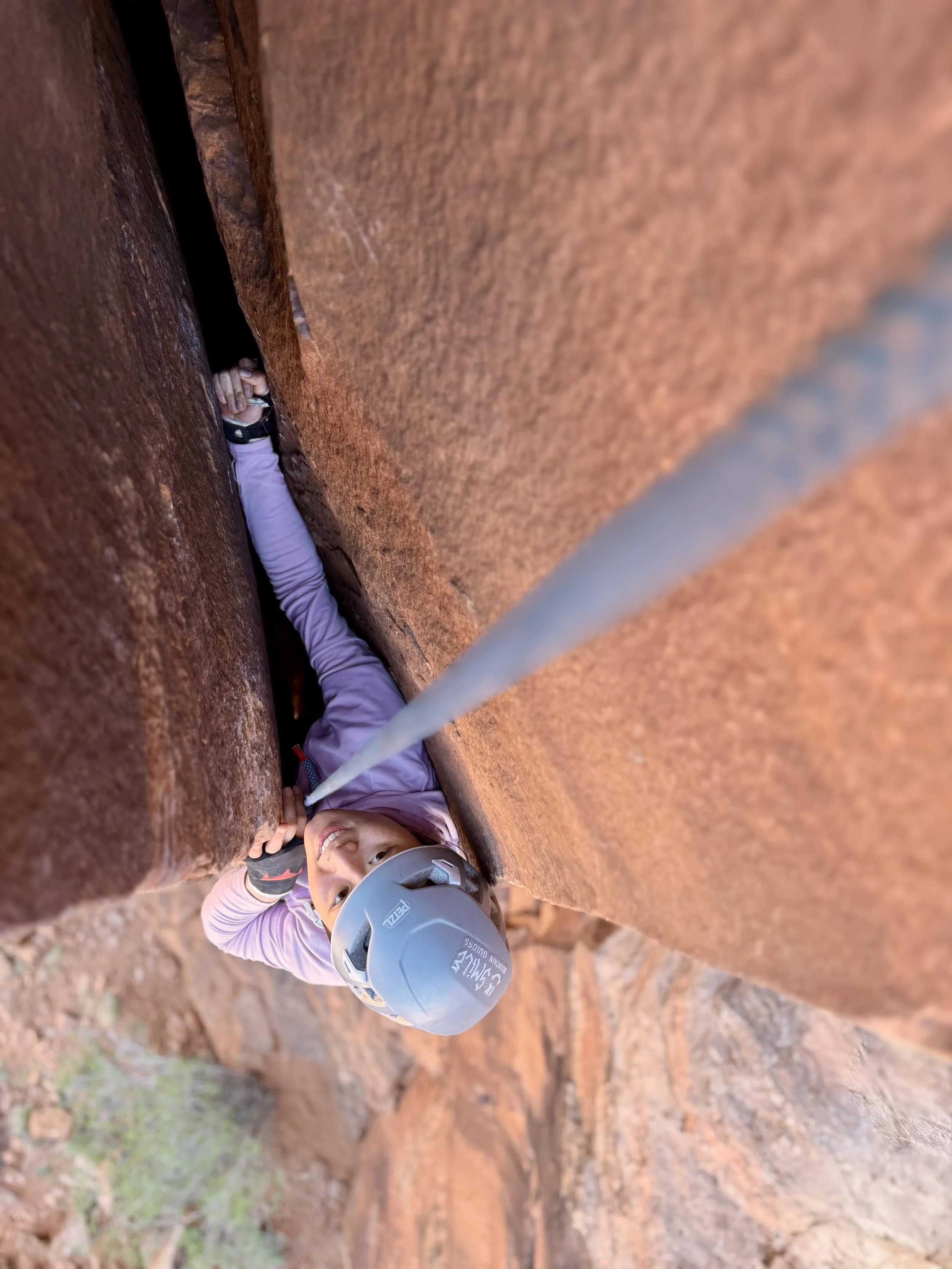 A young woman in a helmet and outdoor clothing climbing a narrow crevice between two large reddish-brown rock walls.