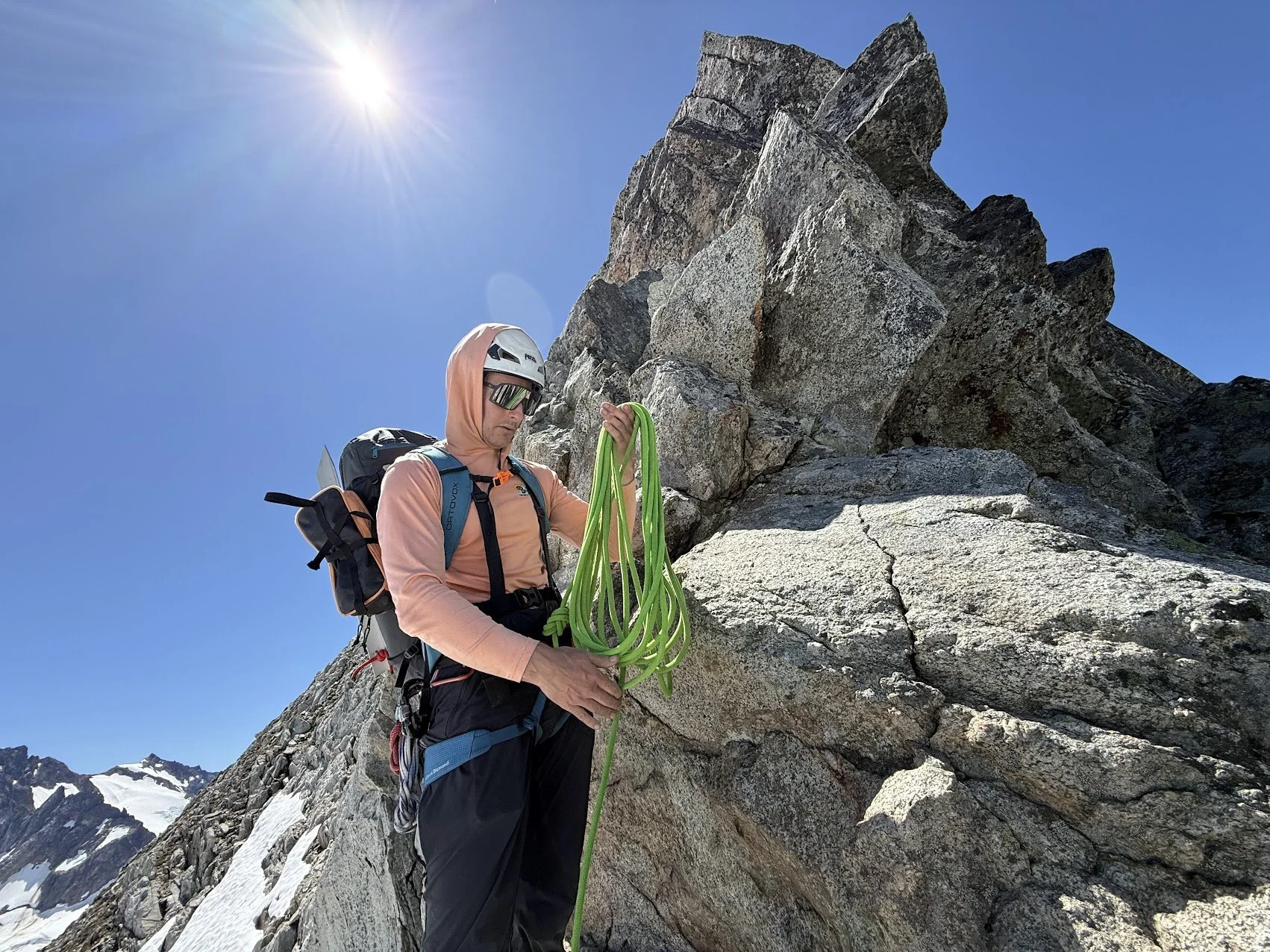 A mountaineer in a helmet and sunglasses attaching a climbing rope to a rock face on a mountain with snow-capped peaks in the background and a clear blue sky.