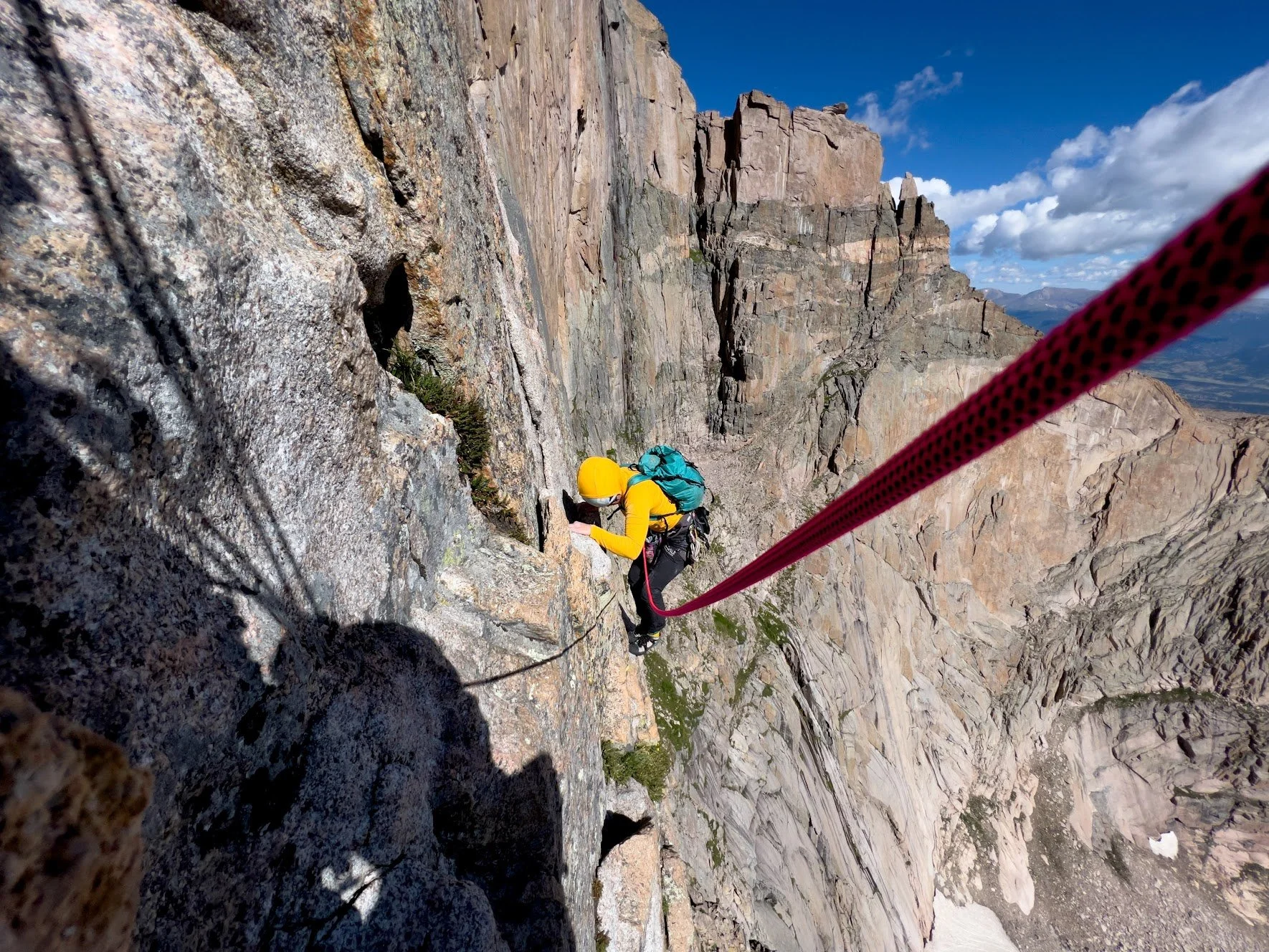 A rock climber in a yellow jacket and helmet ascending a steep cliff with a pink climbing rope in a mountain landscape.