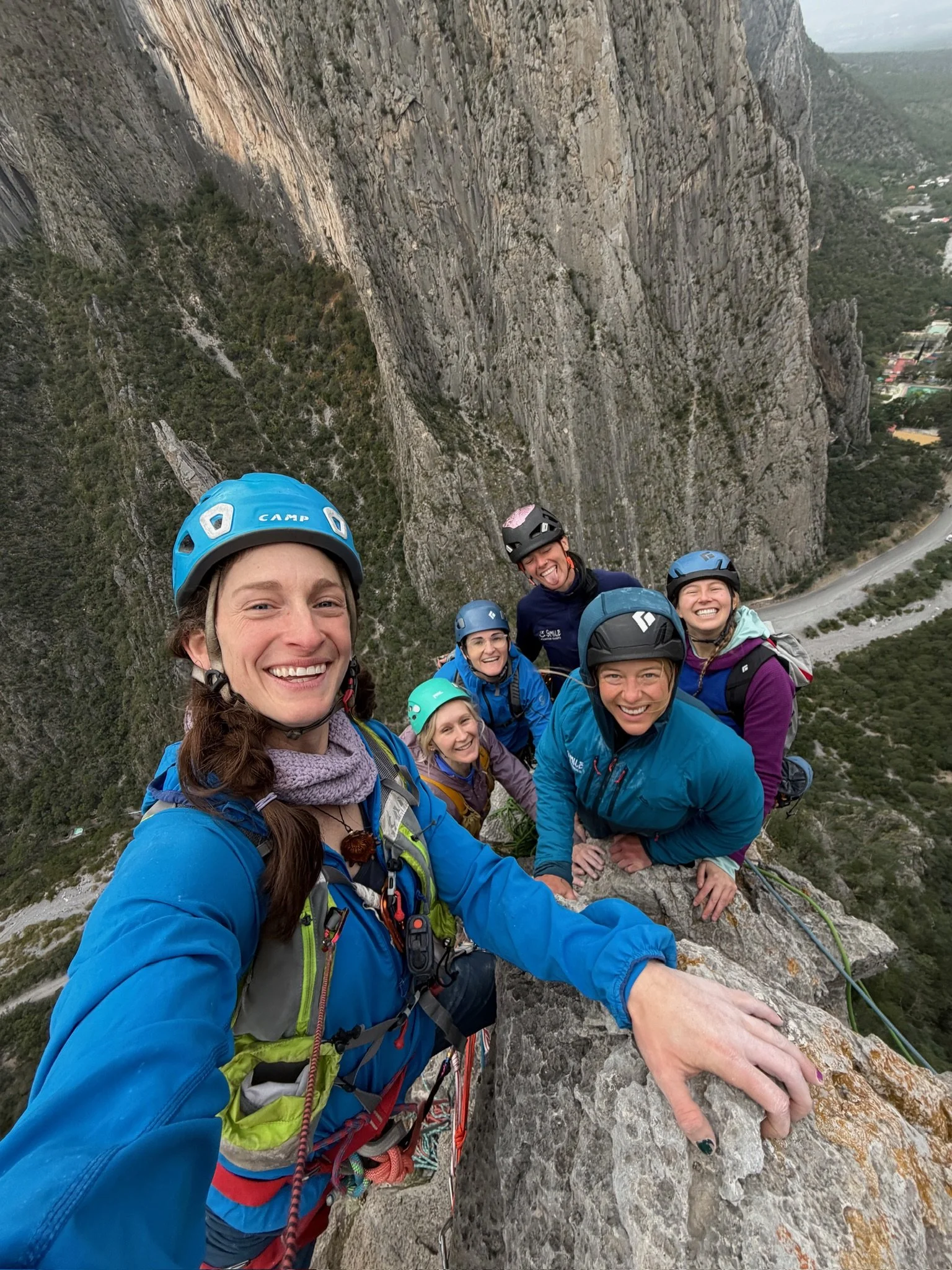Six women in climbing gear, including helmets, smiling at the camera while climbing a steep rock face, overlooking a valley with trees and a road.