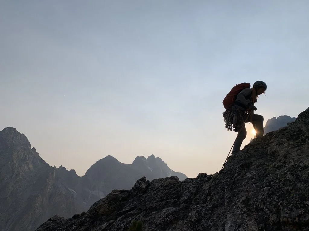 Hiker climbing steep mountain trail with backpack at sunrise or sunset, mountain landscape in background.