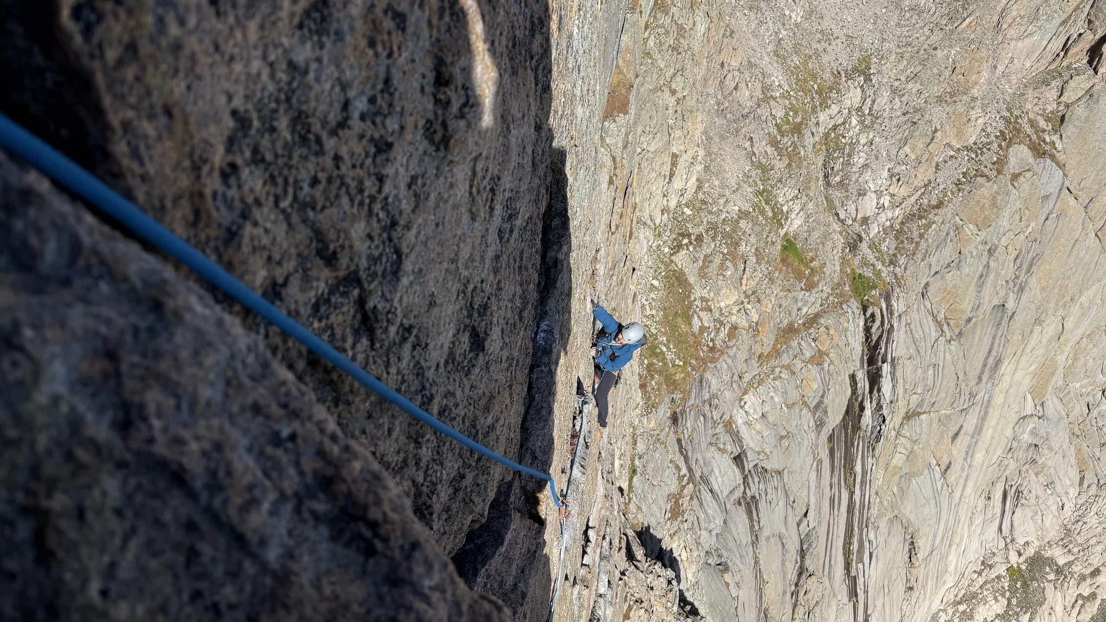 A person is climbing a steep rock face, wearing a helmet and climbing gear, with a safety line attached, on a mountain or rocky terrain.