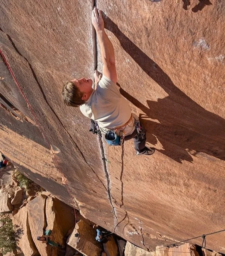 A person climbing a steep rock face outdoors, wearing climbing gear, with view of shadow cast on the rock.