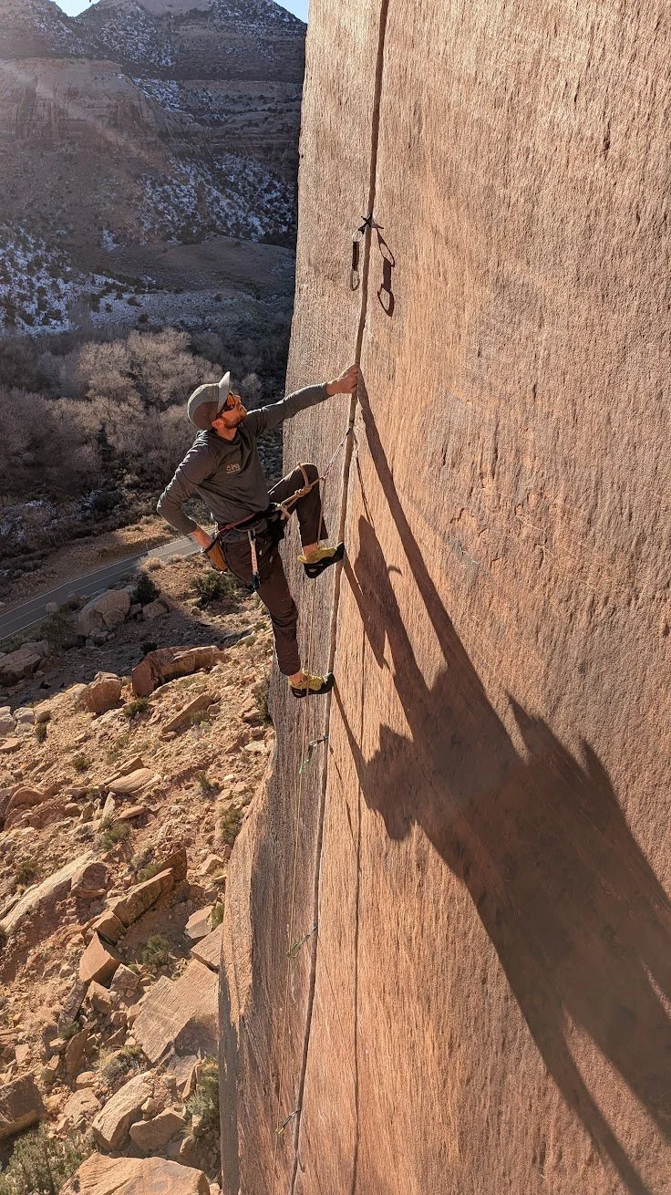 A rock climber ascending a steep red rock face with safety equipment, outdoors in a mountainous area.