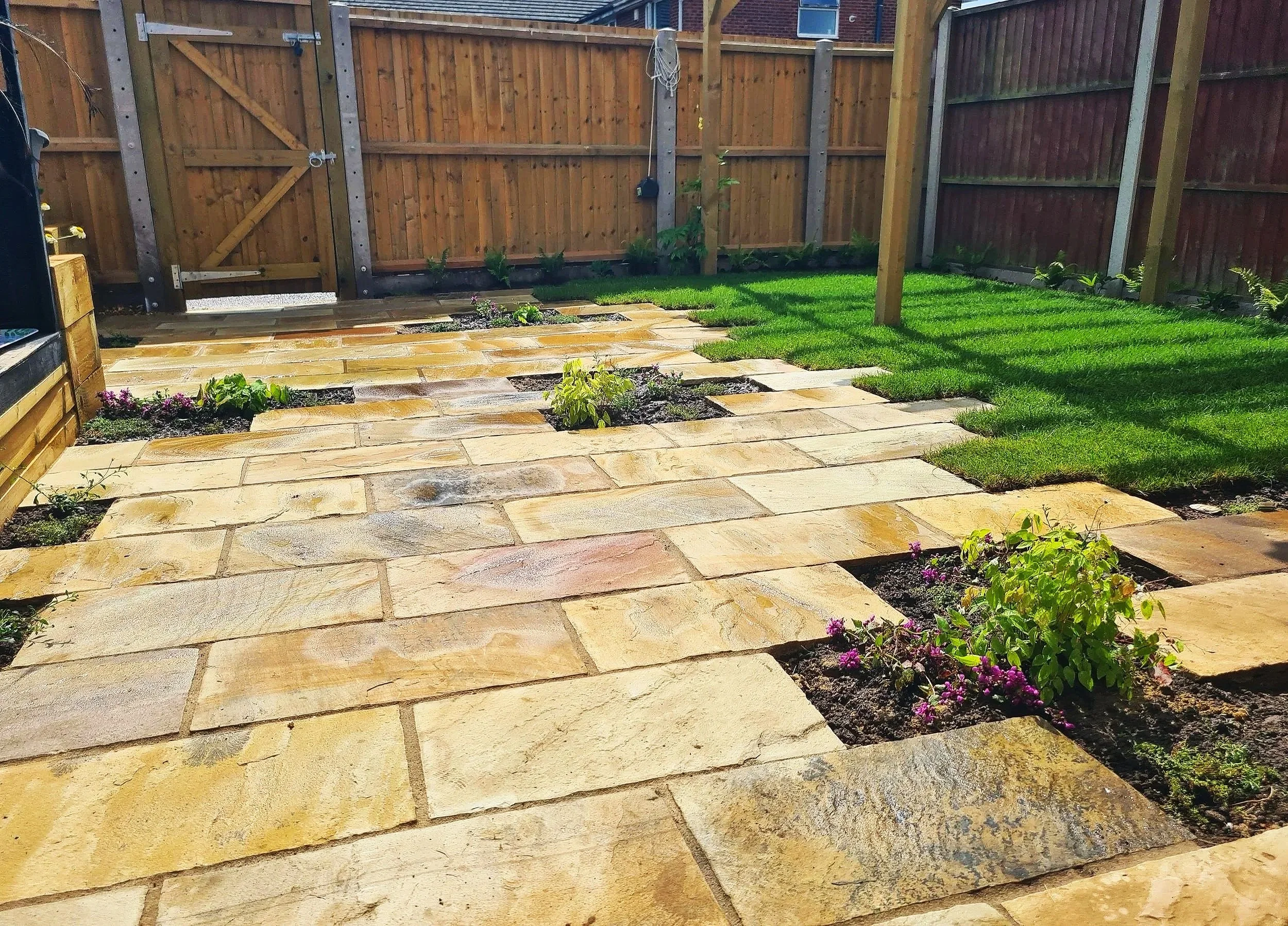 A backyard with a stone patio and flower beds, enclosed by a wooden fence. There is a small patch of grass and a post in the yard.