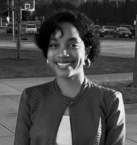 A woman smiling at night by a river with city skyline lights reflecting on the water, wearing glasses and a knit winter hat with a pom-pom.