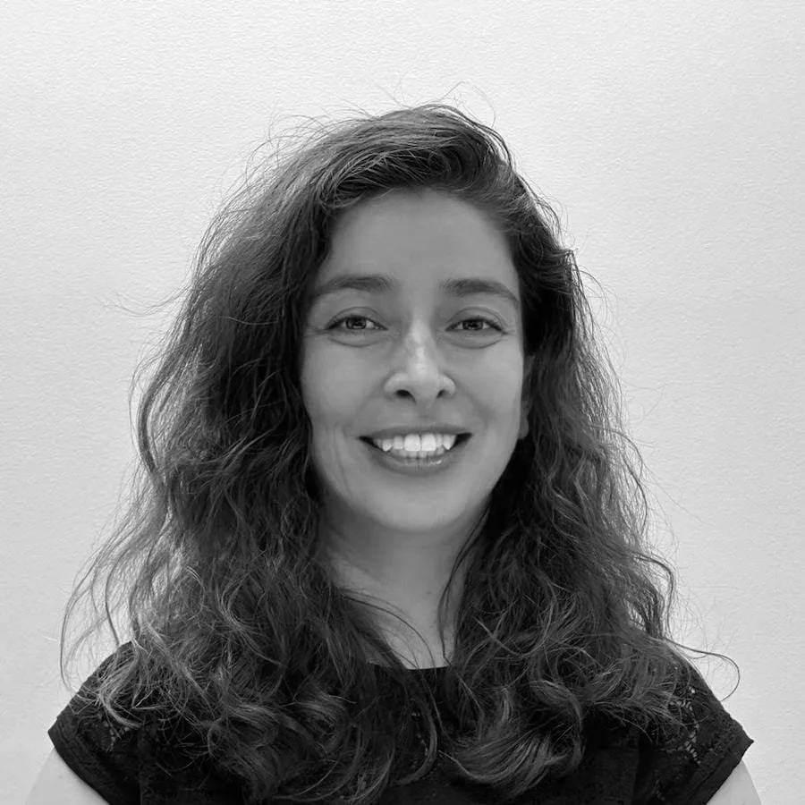 Black and white portrait of a woman with long hair wearing a sleeveless top and earrings. She is looking towards the camera against a plain background.