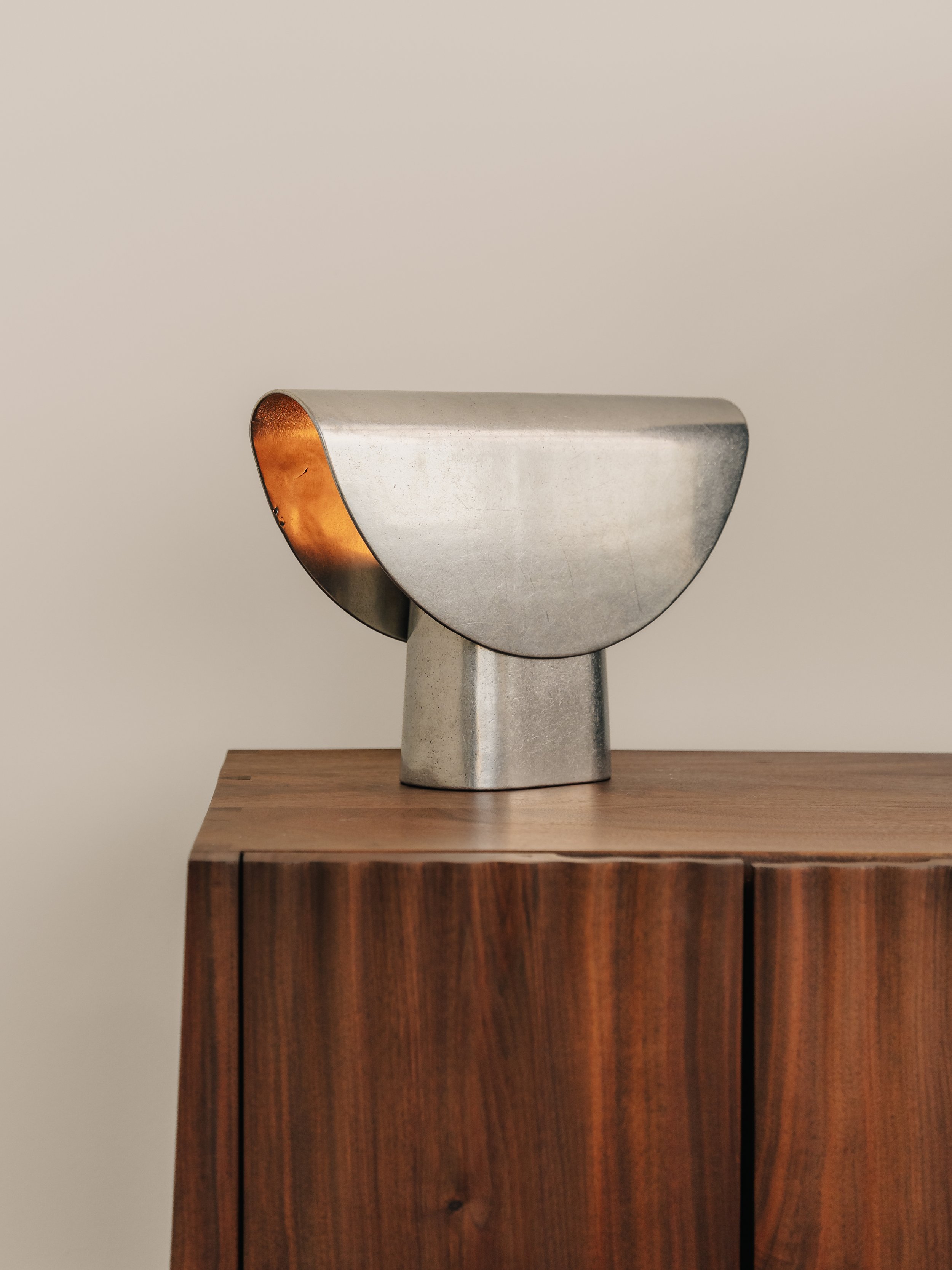 A modern metallic sculpture with an abstract design, placed on top of a wooden cabinet with a dark finish, against a neutral wall.